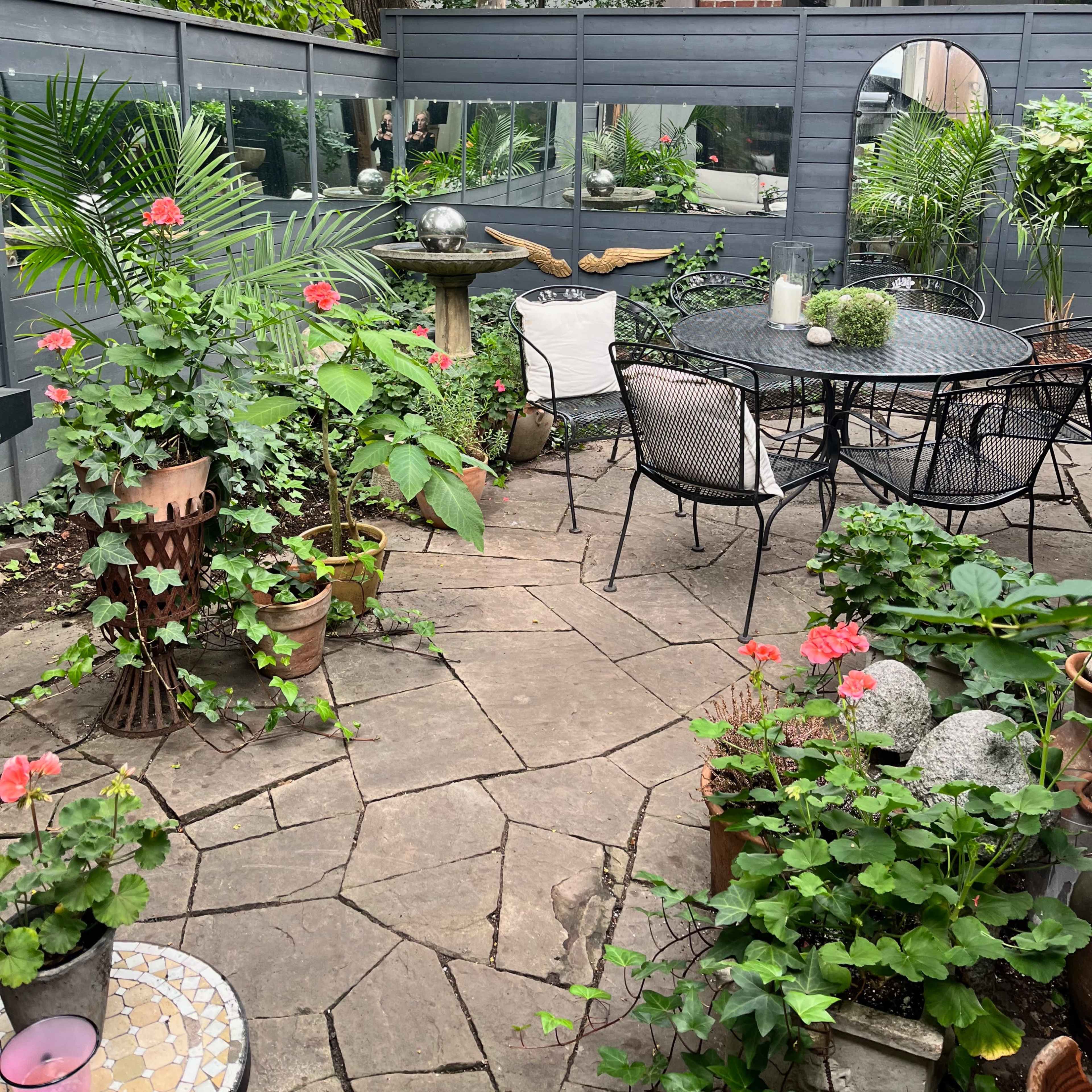 A garden patio features a circular table surrounded by chairs, stone pathways, and diverse potted plants, with a background of a brick building.