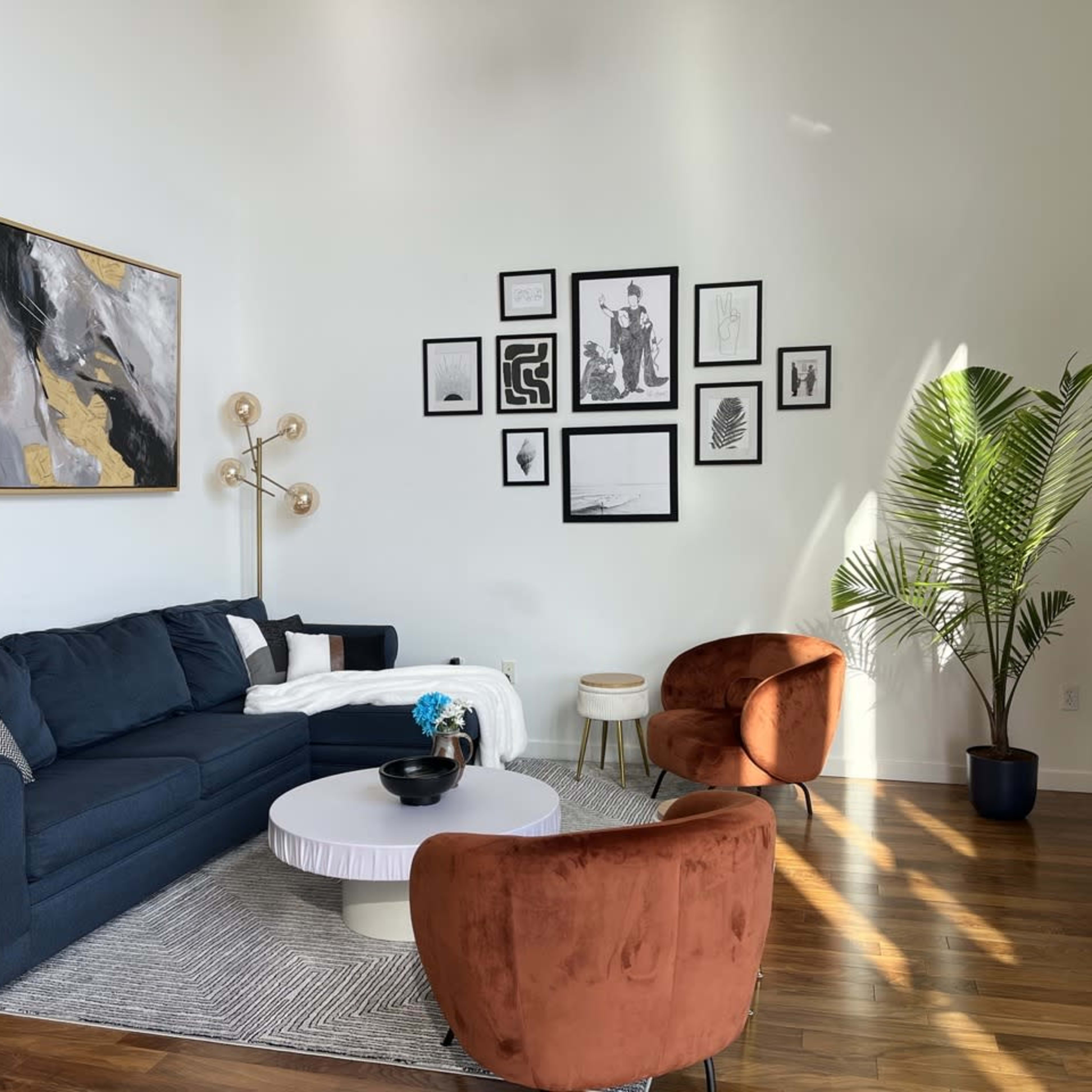 A modern living room features a dark blue sofa, two orange armchairs, a round white coffee table, and a wall adorned with black and white framed artwork, alongside a tall green plant.
