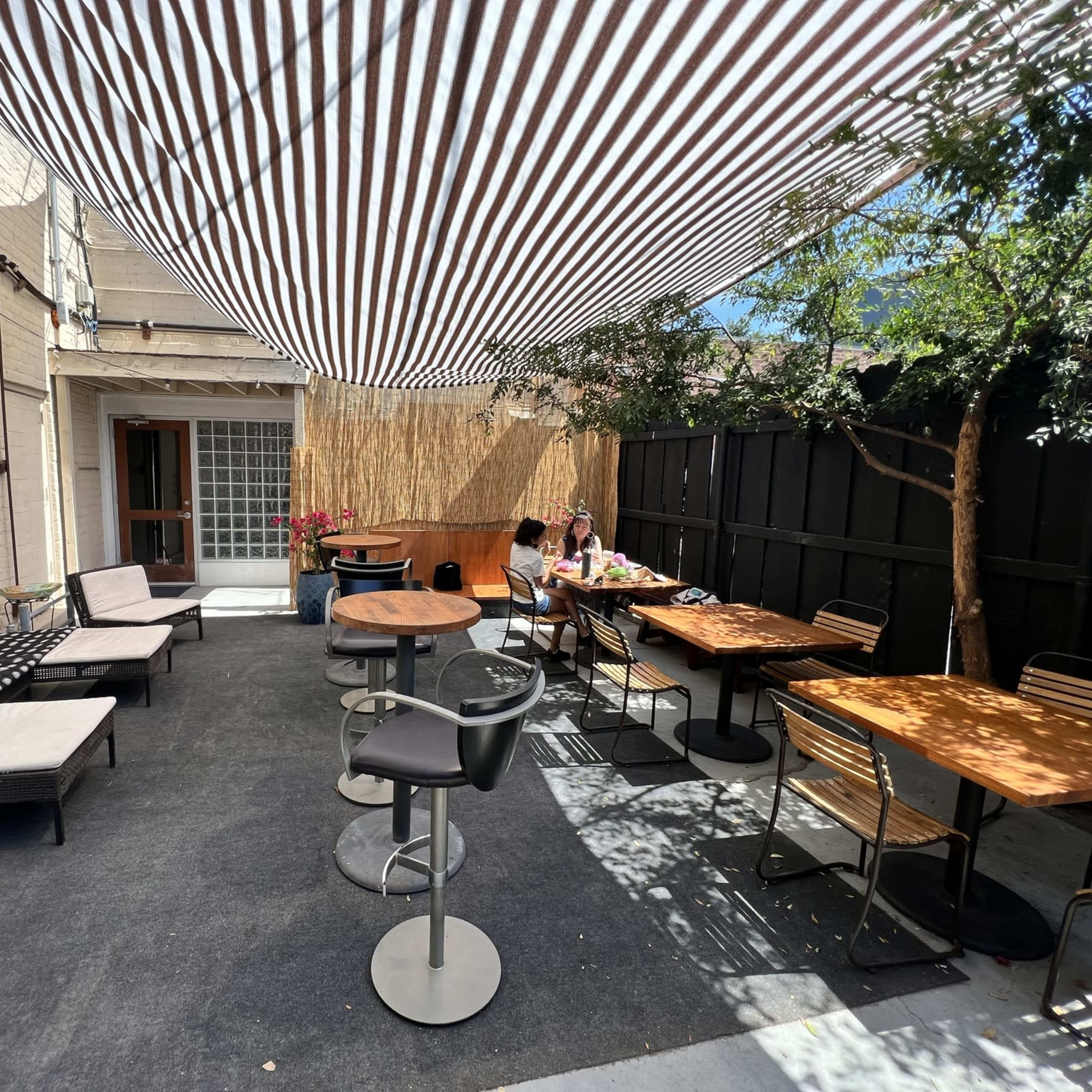 A backyard patio area with striped overhead shading, tables, and seating arrangements, where two people are sitting at a table.