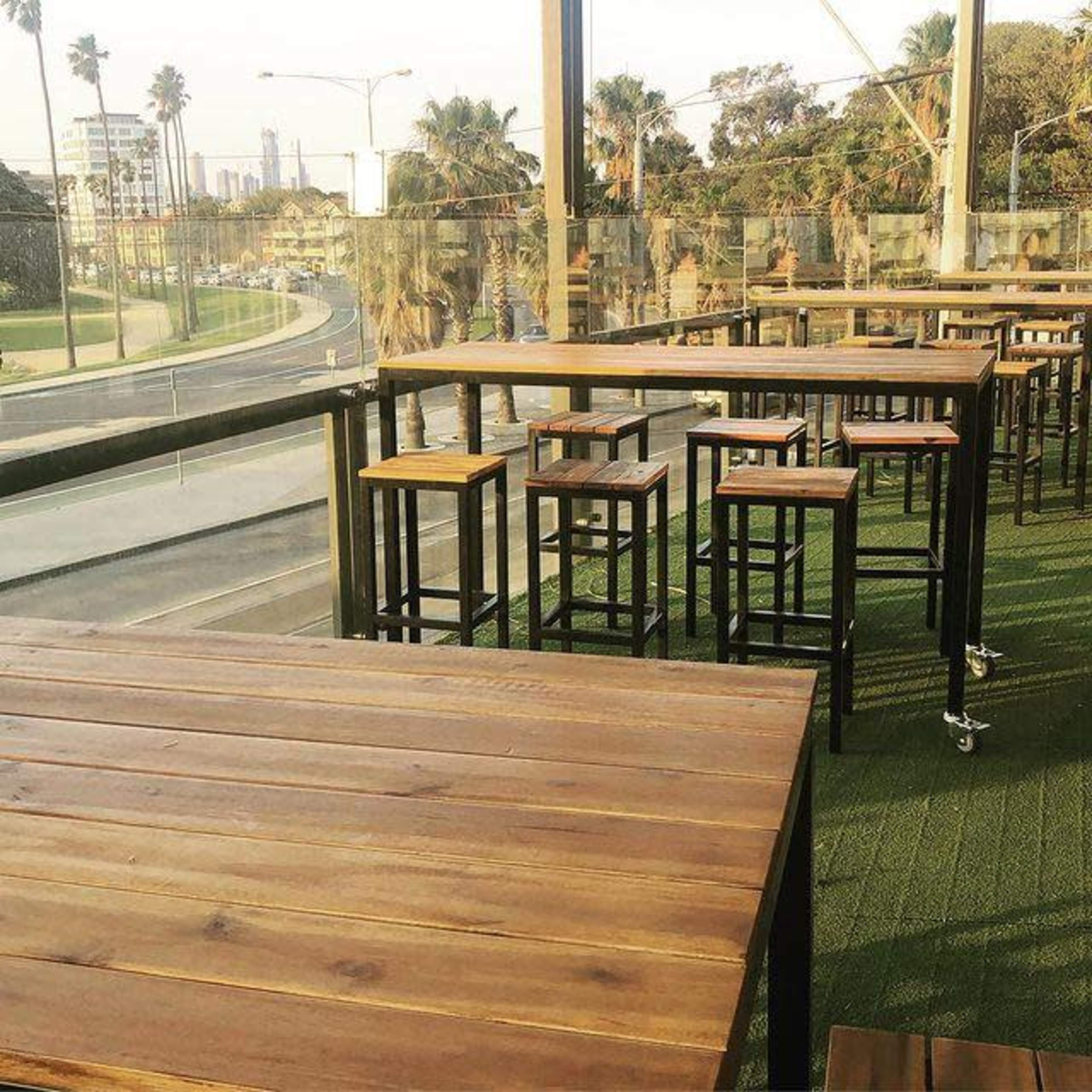 A wooden patio area with tall tables and stools overlooks a road lined with palm trees.