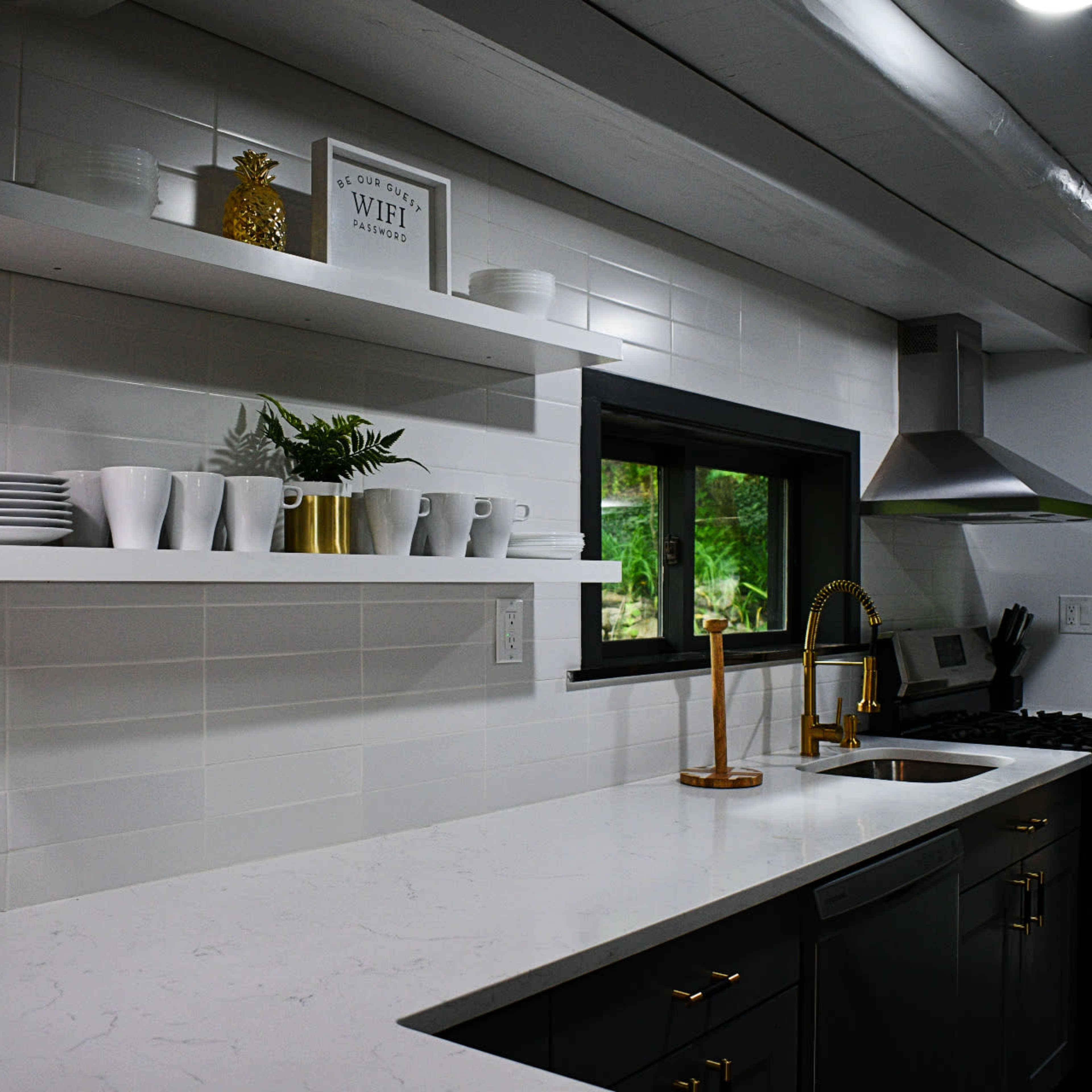A modern kitchen with white tile walls, a large countertop, open shelves displaying dishware, and a view of an outdoor stone area through a glass door.