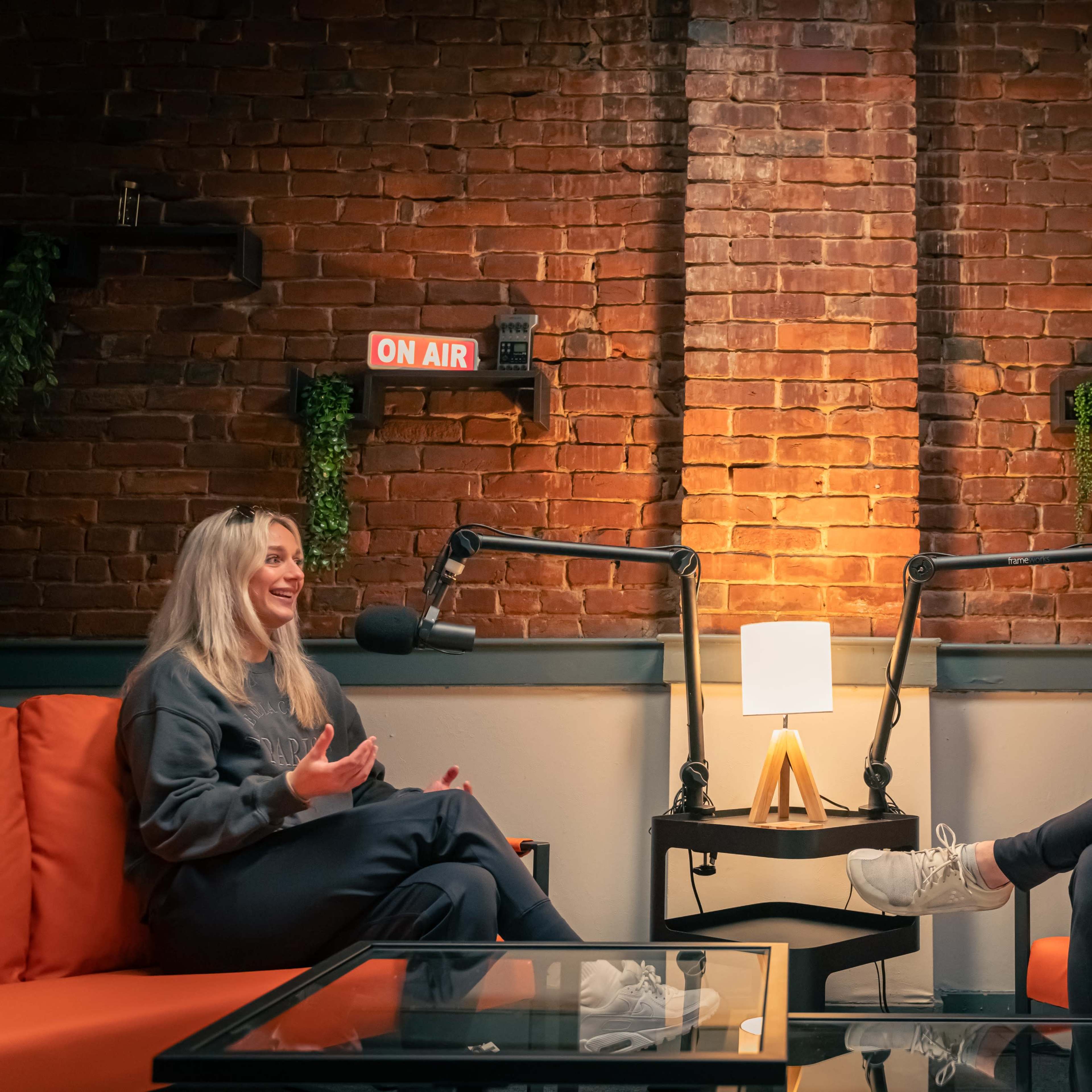 Two individuals are seated in a cozy studio setting with orange couches, engaged in conversation while microphones are positioned in front of them, and a lamp illuminates the space.