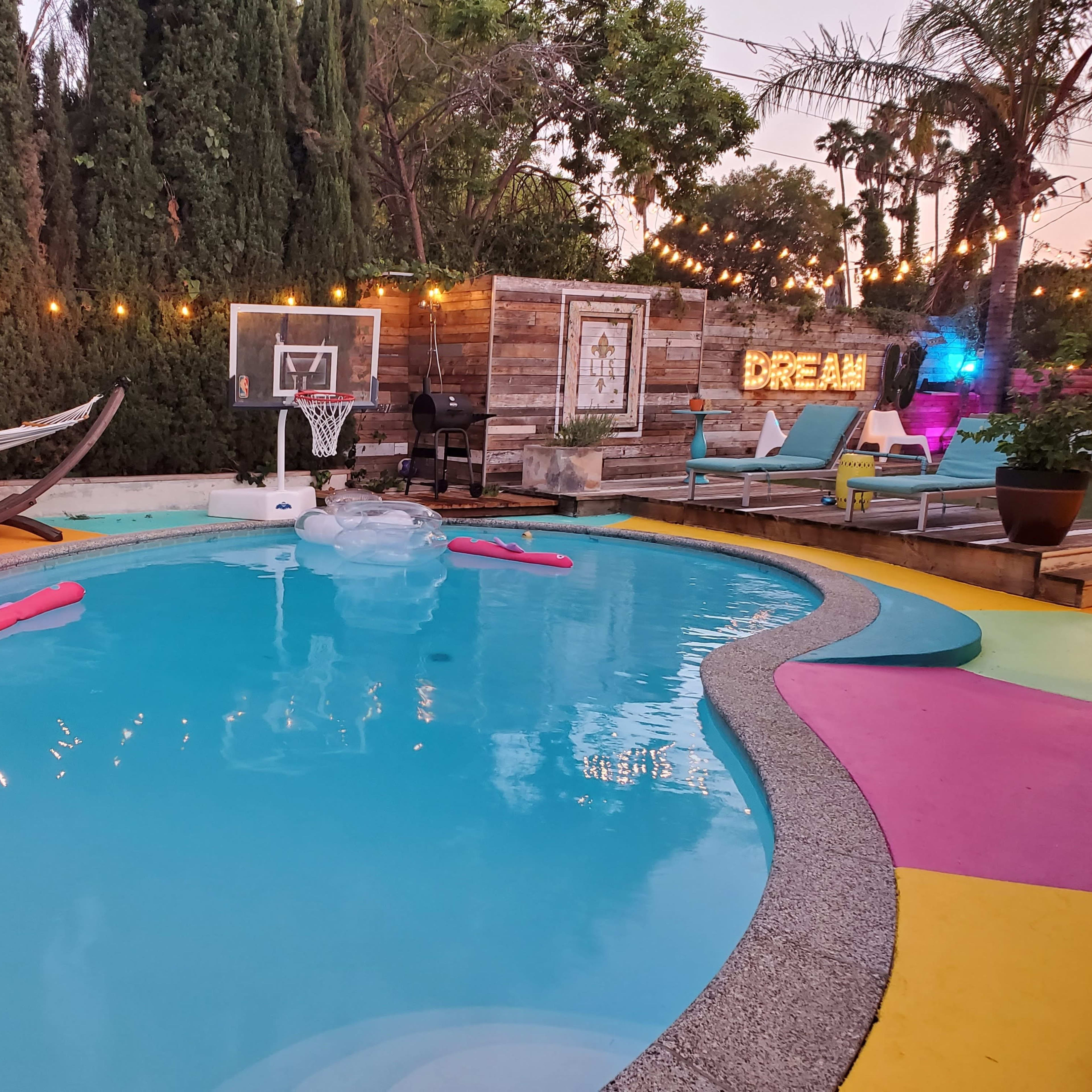 The scene features a swimming pool surrounded by colorful decking, a basketball hoop, a hammock, and illuminated decorations in a landscaped backyard.