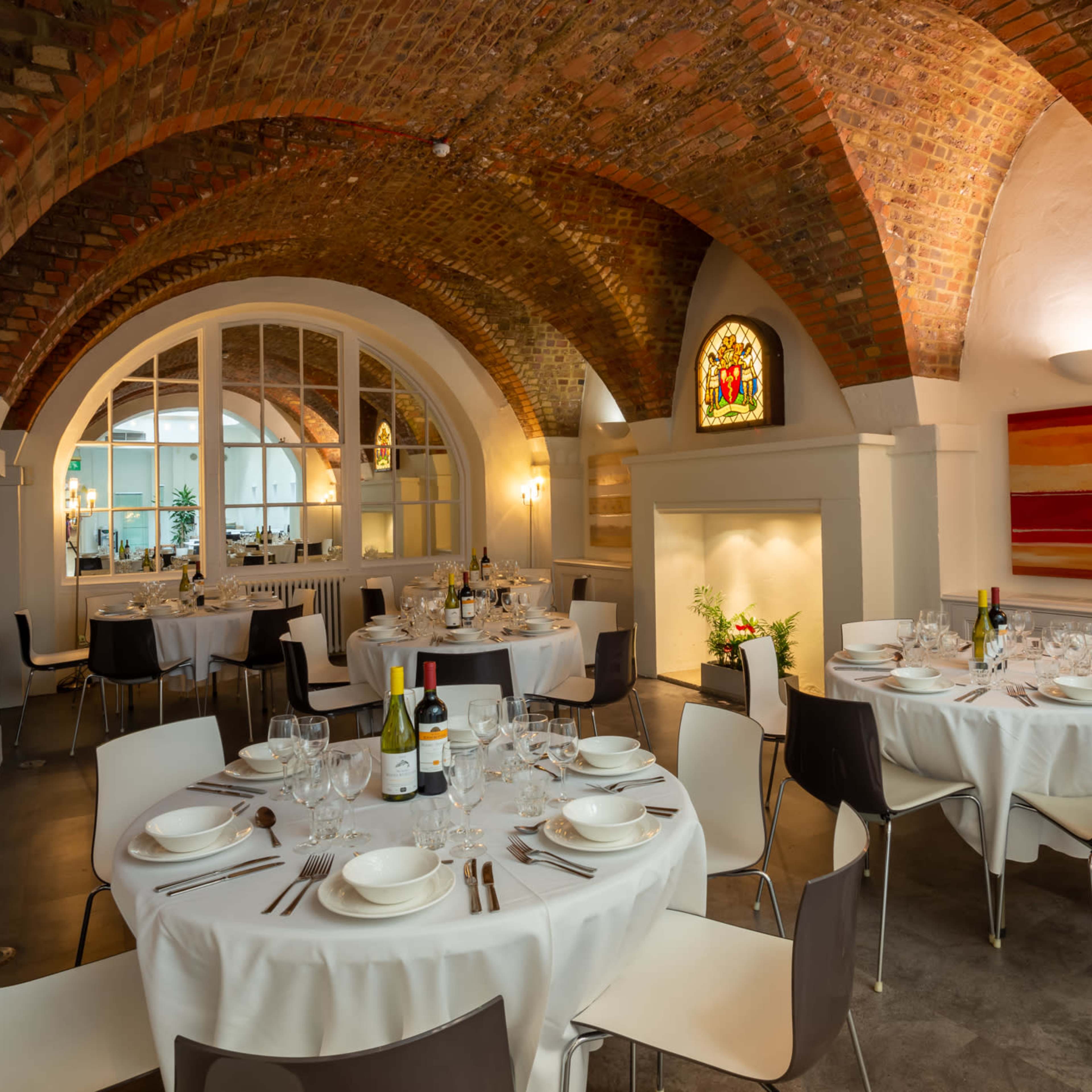 A dining area in a restaurant with arched brick ceilings, round tables set with white tablecloths, and large windows letting in light.