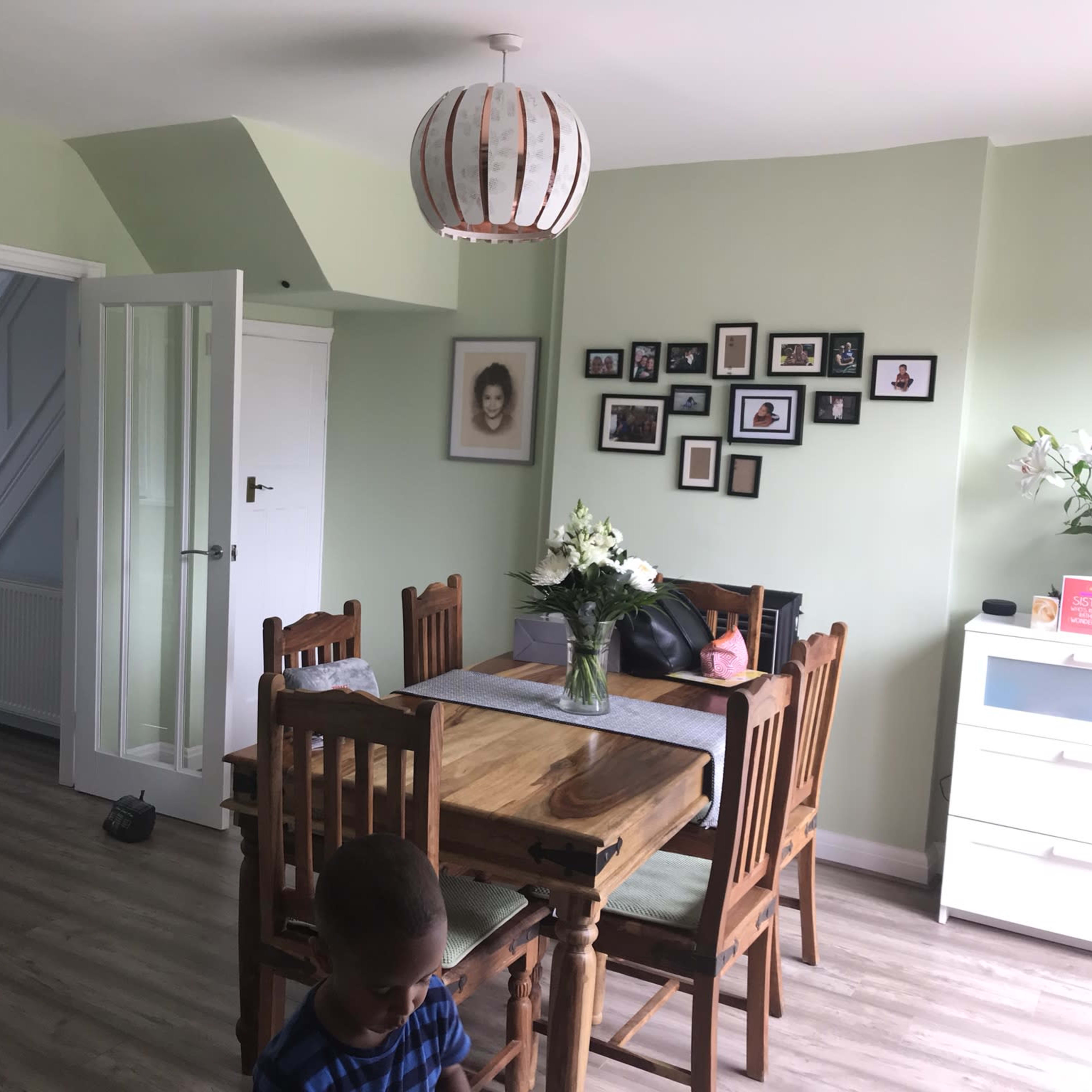 A dining area features a wooden table surrounded by chairs, a flower vase, and walls adorned with pictures, while an adjacent room can be seen through an open door.