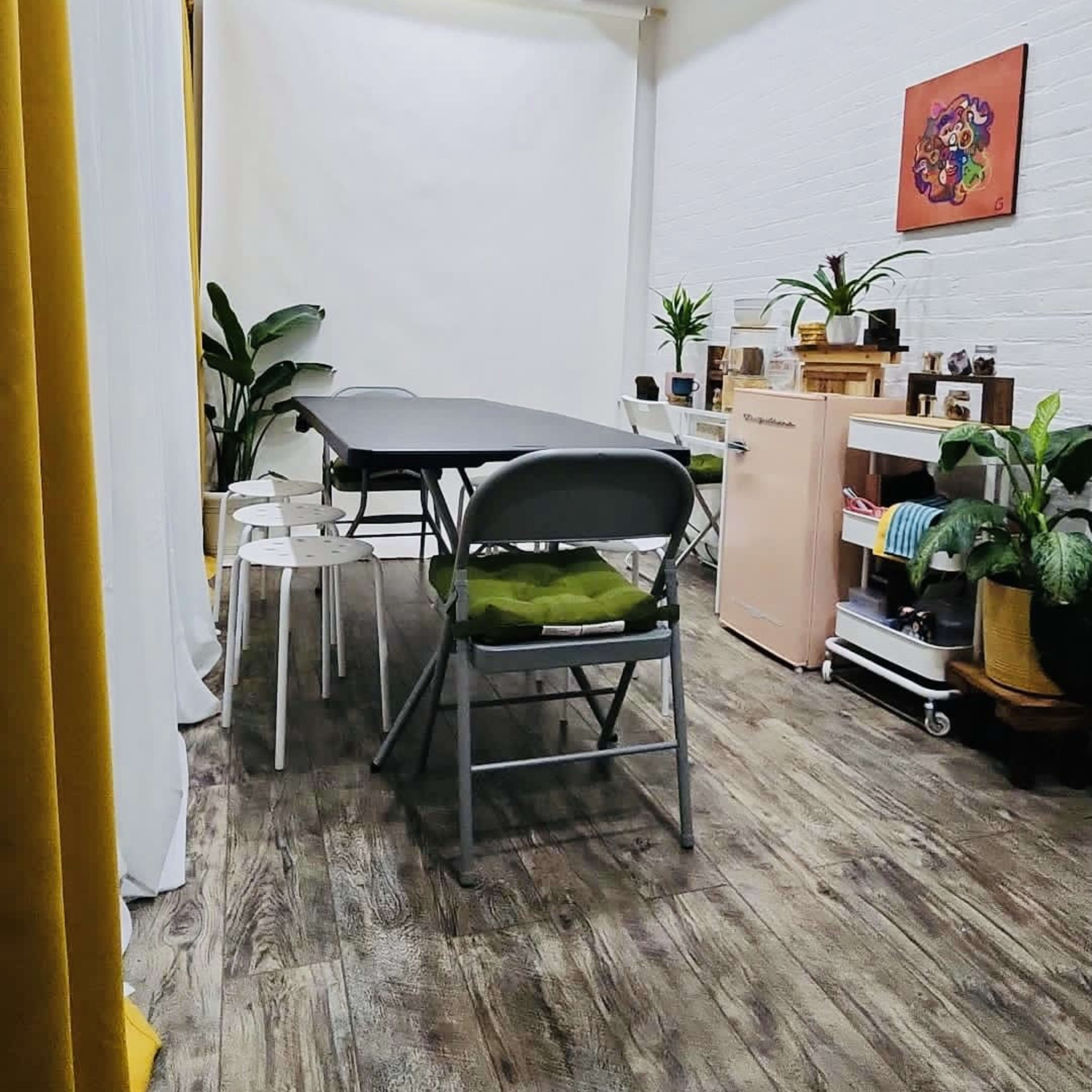 A bright, organized room with a long table, folding chairs, plants, and shelves lined with various items.