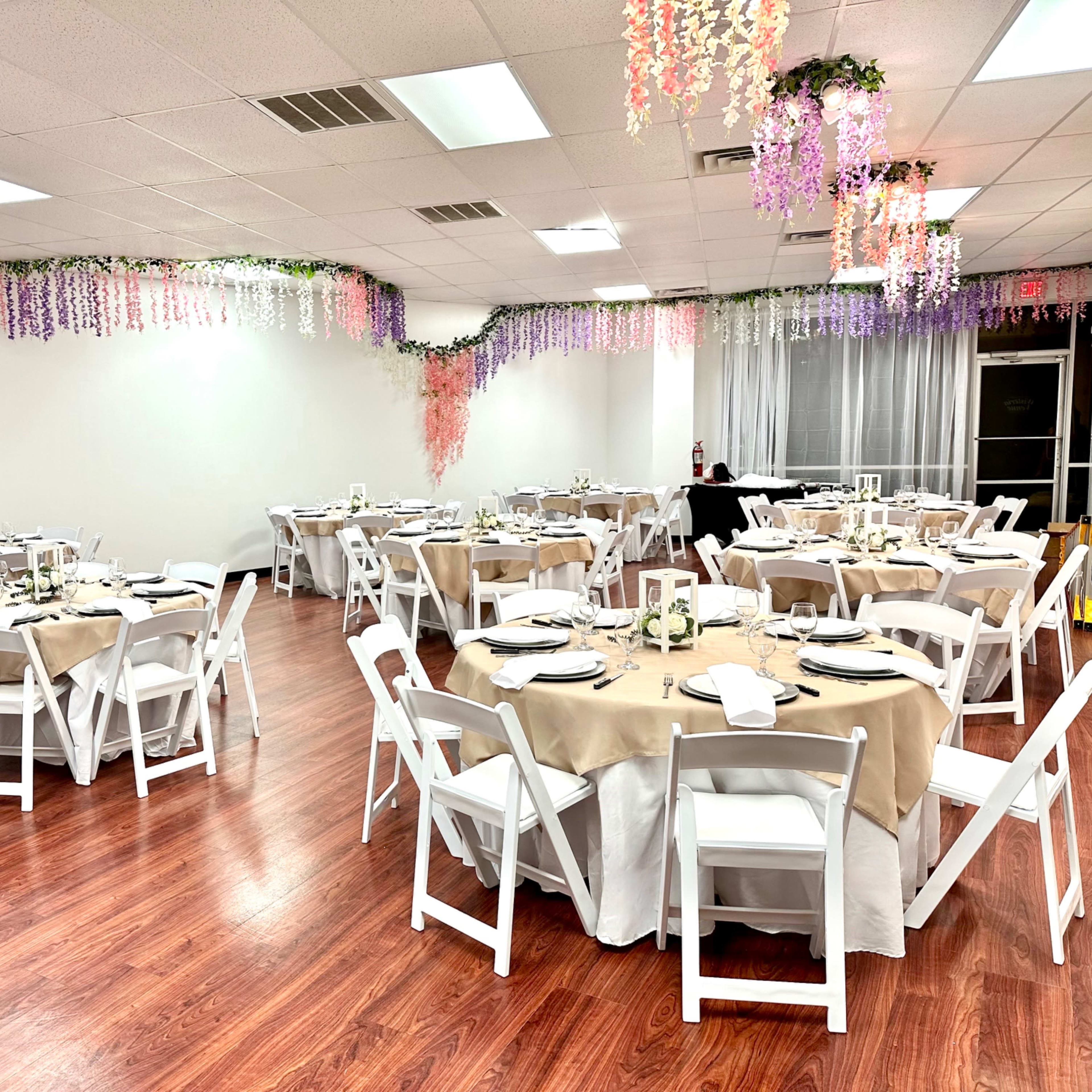 A banquet hall features several round tables covered with beige tablecloths, surrounded by white folding chairs, and adorned with colorful decorations hanging from the ceiling.