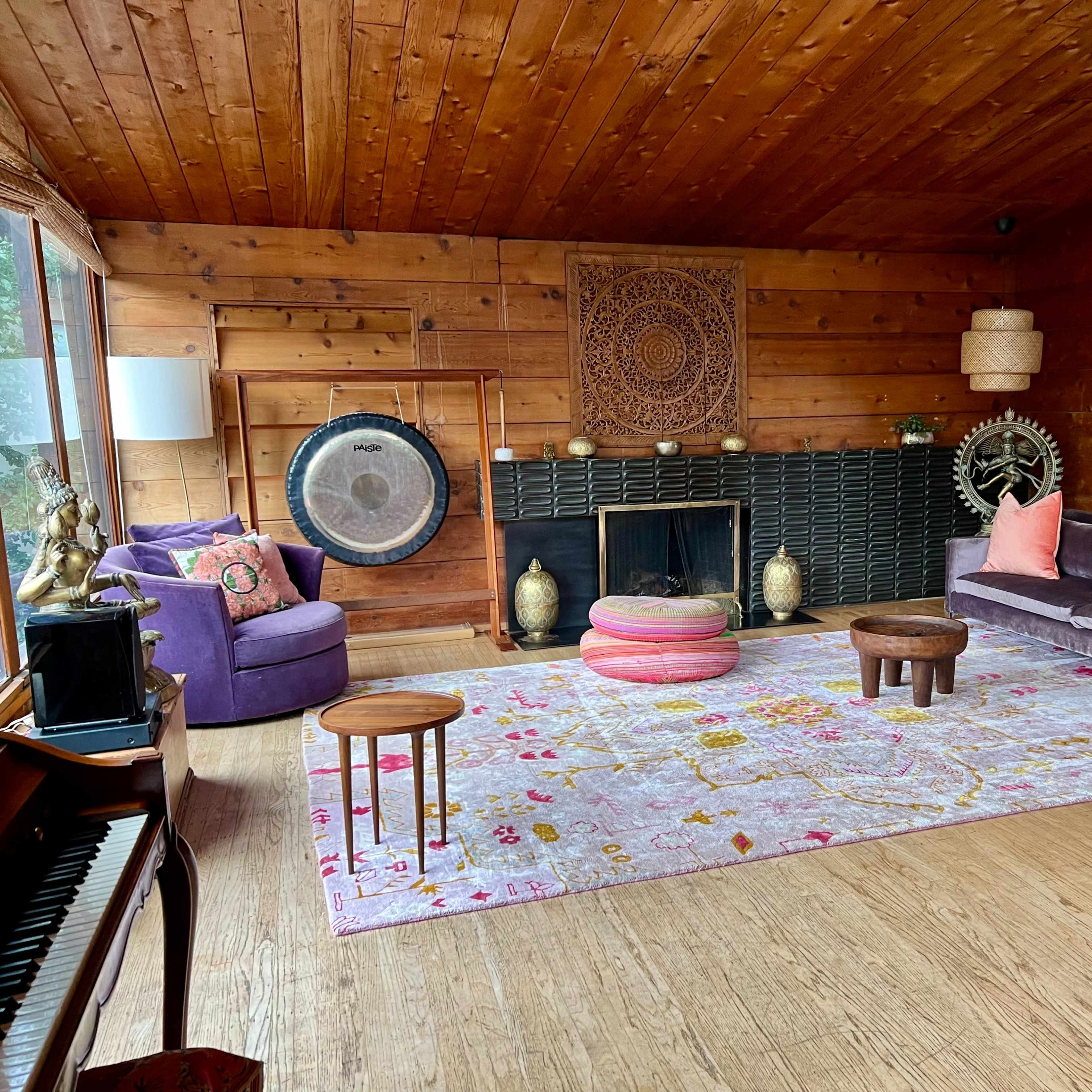 A well-decorated living room with wooden walls, a large pink area rug, a fireplace, and various decorative elements including a gong and a large mirror.