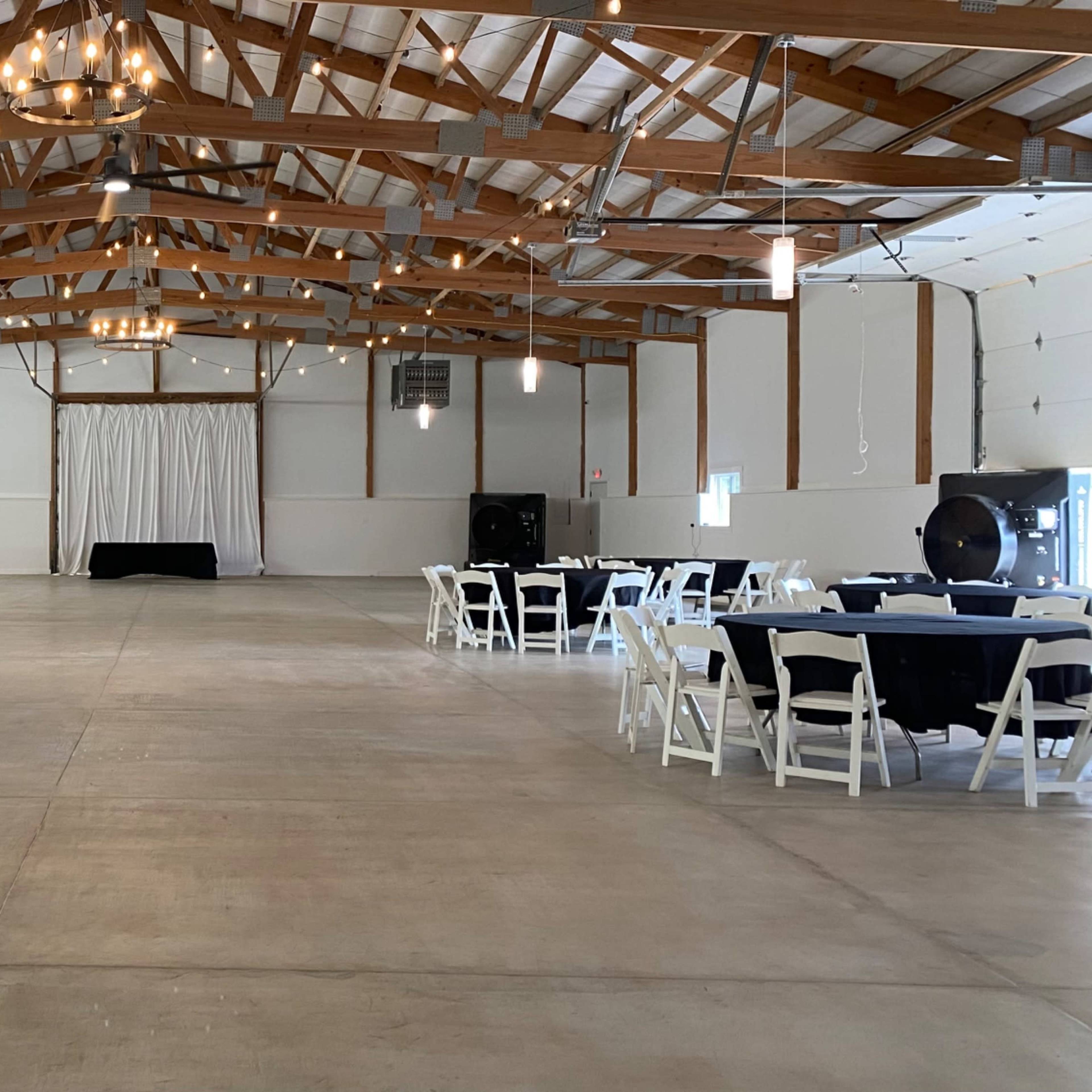 A spacious hall with wooden beams, featuring several round tables set with white tablecloths and black accents, and a stage area at the front.
