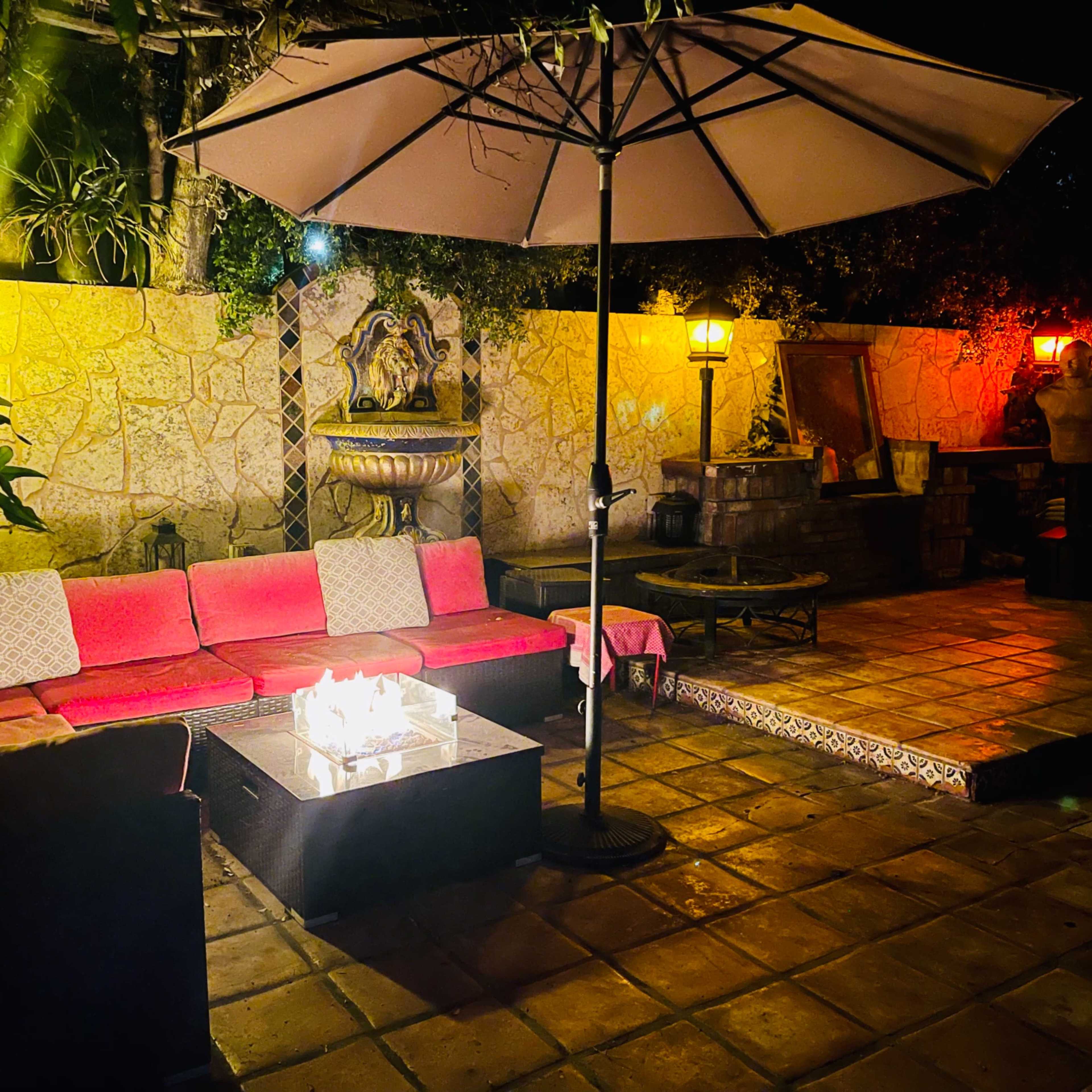 A cozy outdoor seating area with a pink sofa, a fire pit, and an umbrella, illuminated by wall lamps at night.