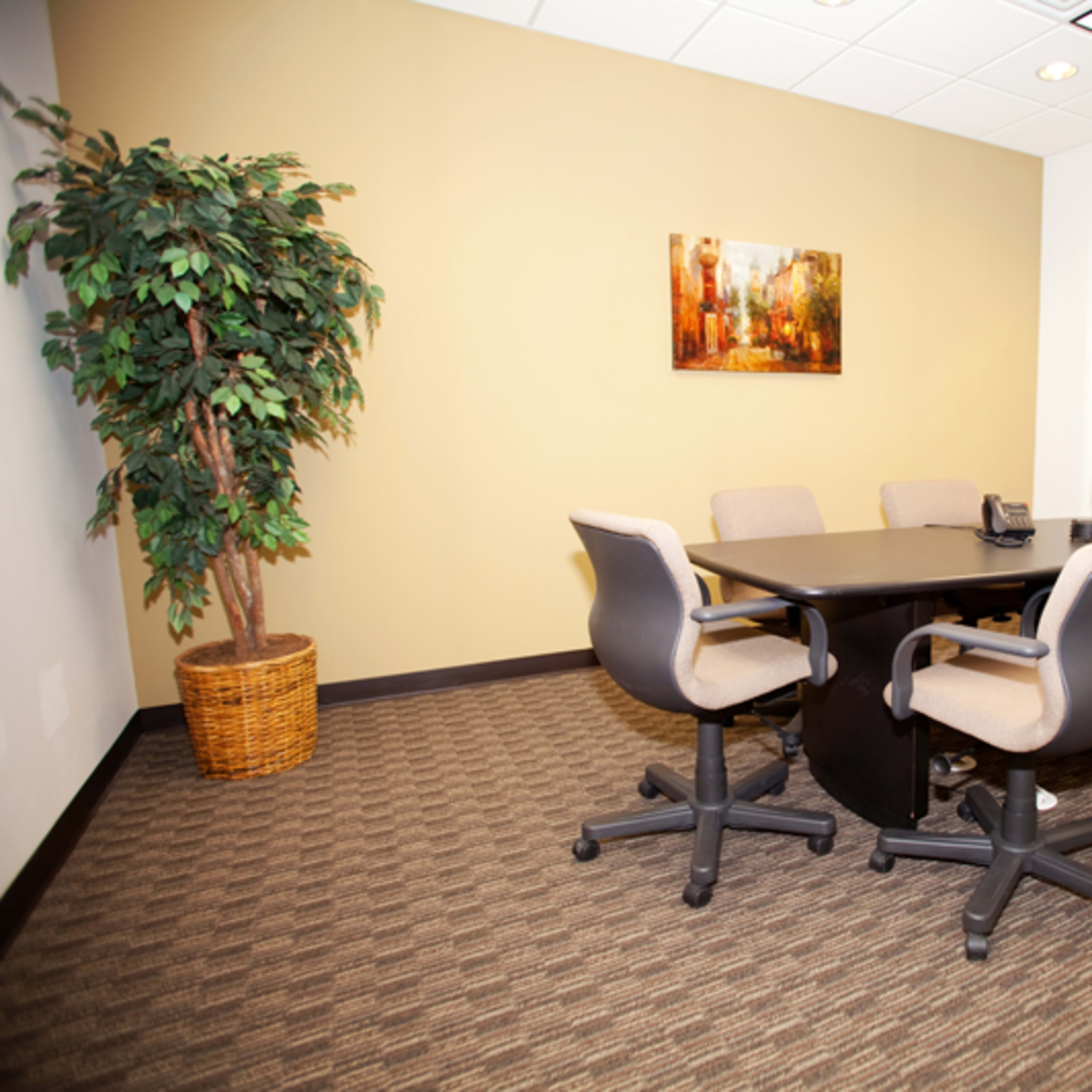 A conference room with a round black table surrounded by five wheeled office chairs, a potted plant in the corner, and a framed painting on the wall.