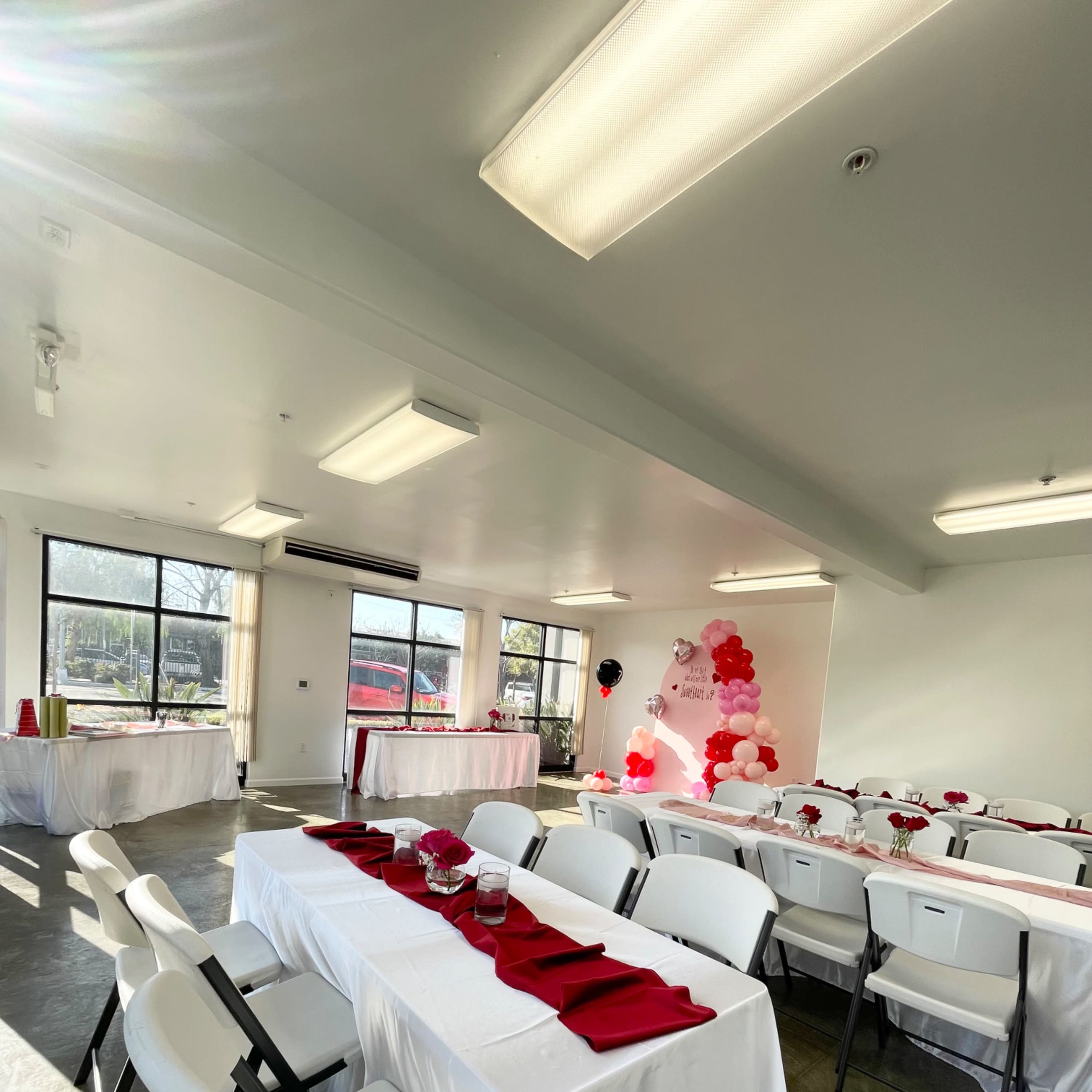 A banquet room set up with white tables covered in tablecloths, red napkins placed neatly on each setting, and a balloon decoration in the corner, all illuminated by natural light from large windows.