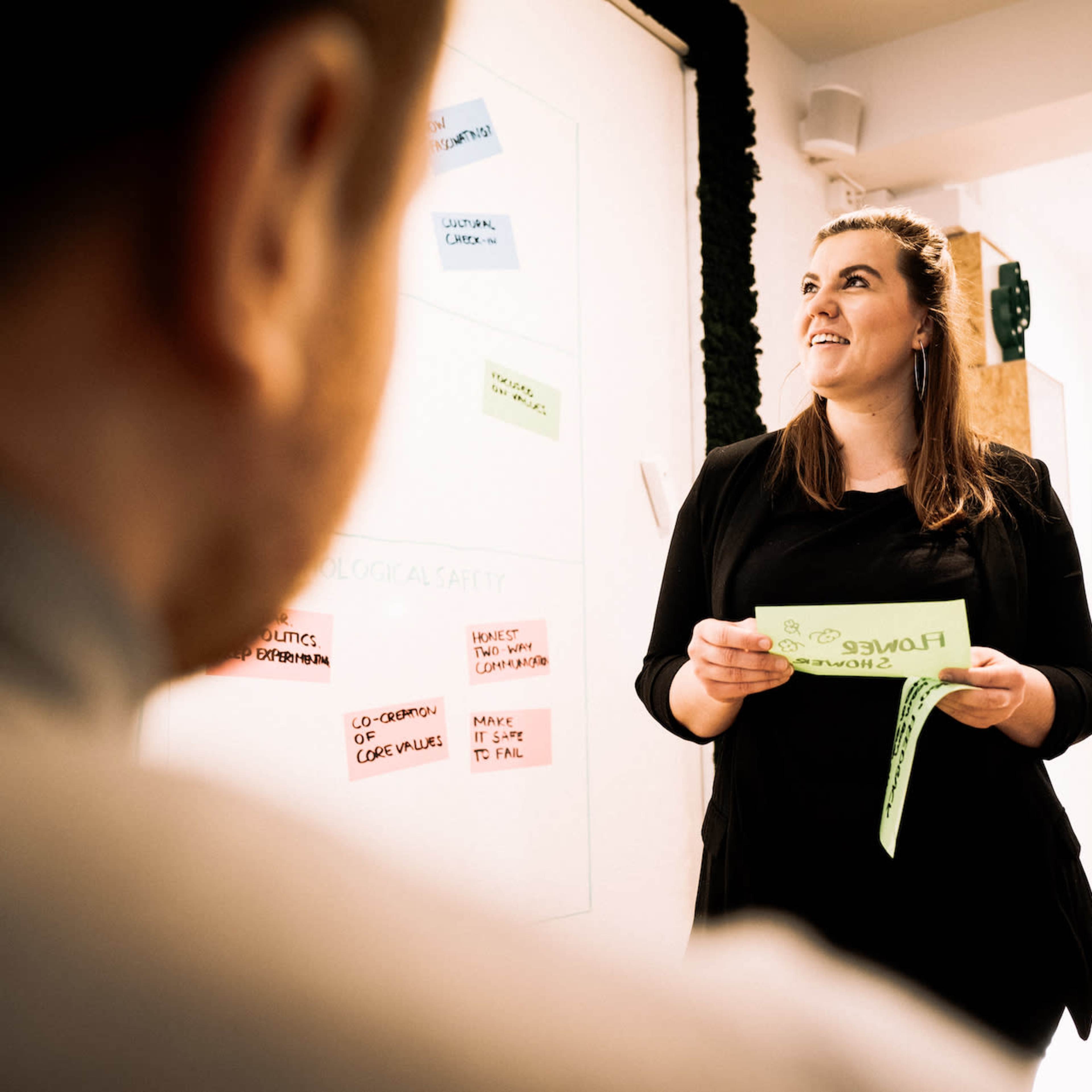 A woman stands in front of a whiteboard displaying colorful sticky notes while holding a green paper, speaking to a group.