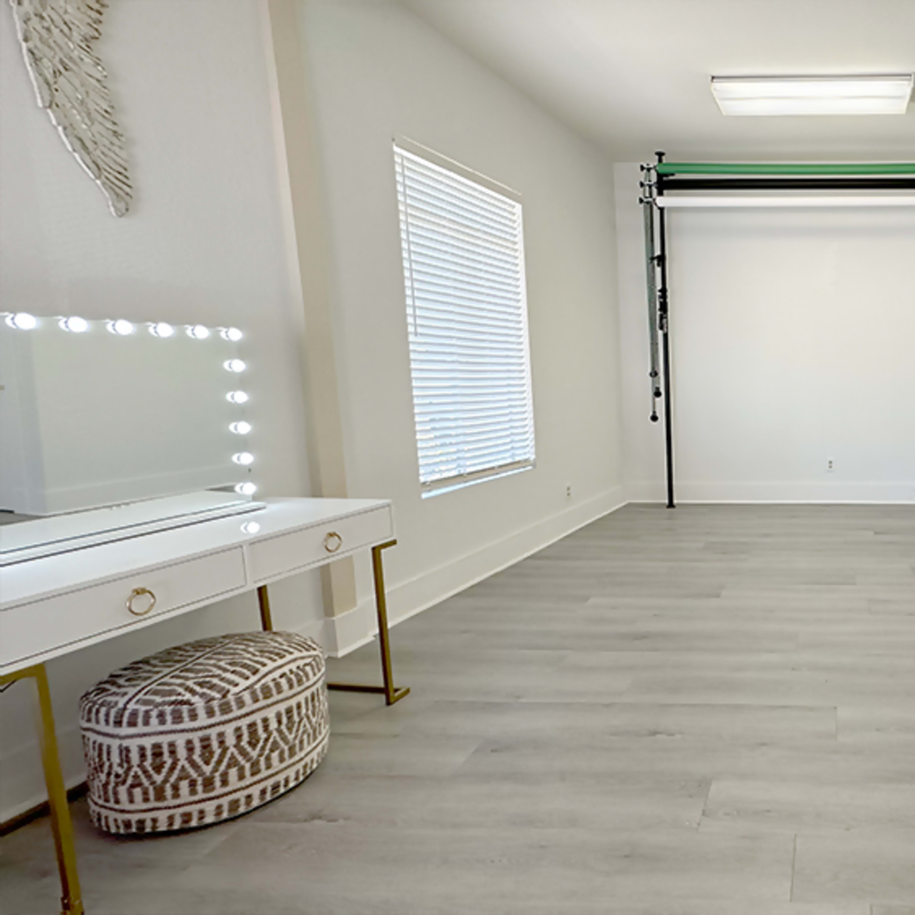 A spacious room features a large window, a folding backdrop stand, and a vanity with illuminated mirrors.