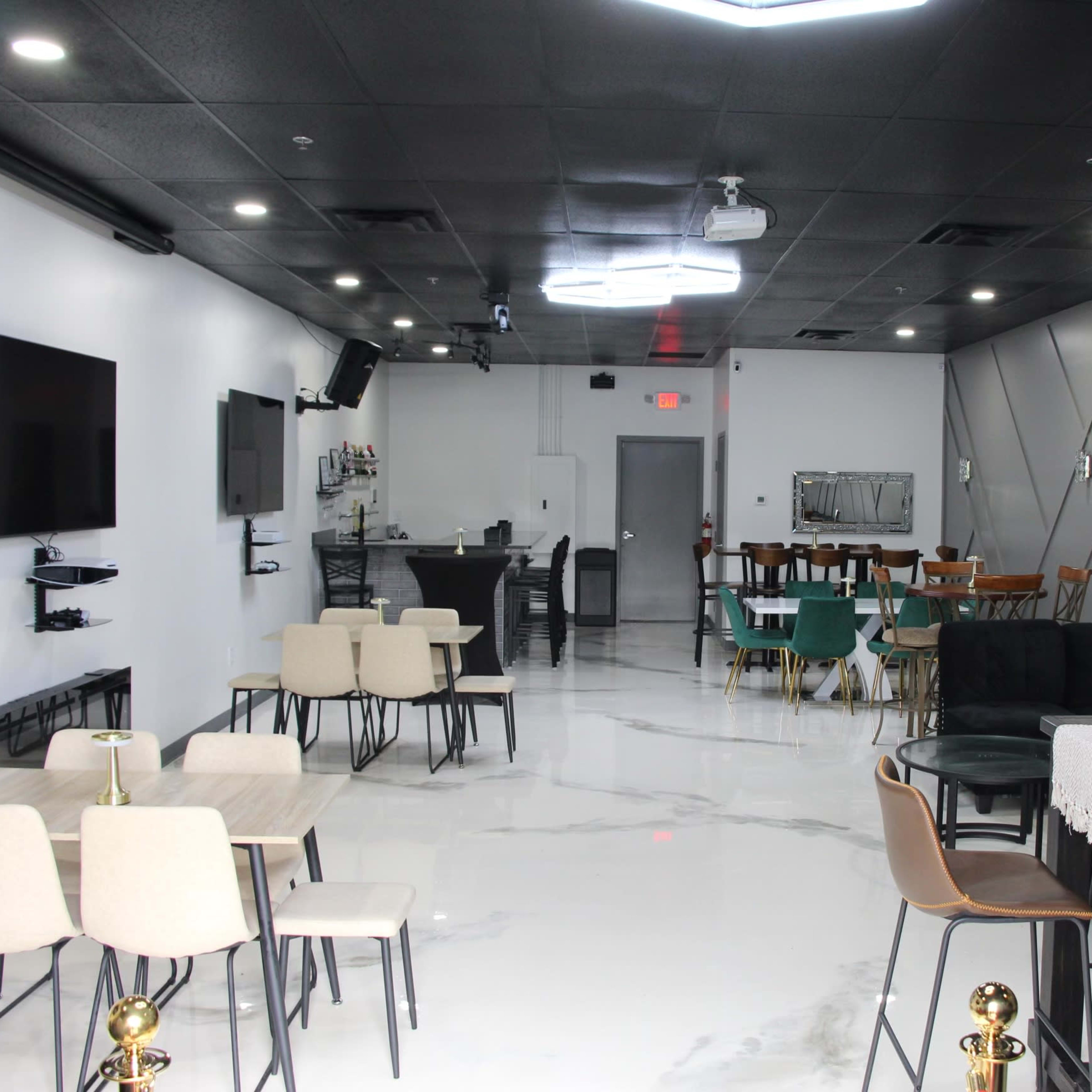 A modern lounge area featuring several tables and chairs, a bar setup, and wall-mounted televisions in a well-lit space with a polished floor.