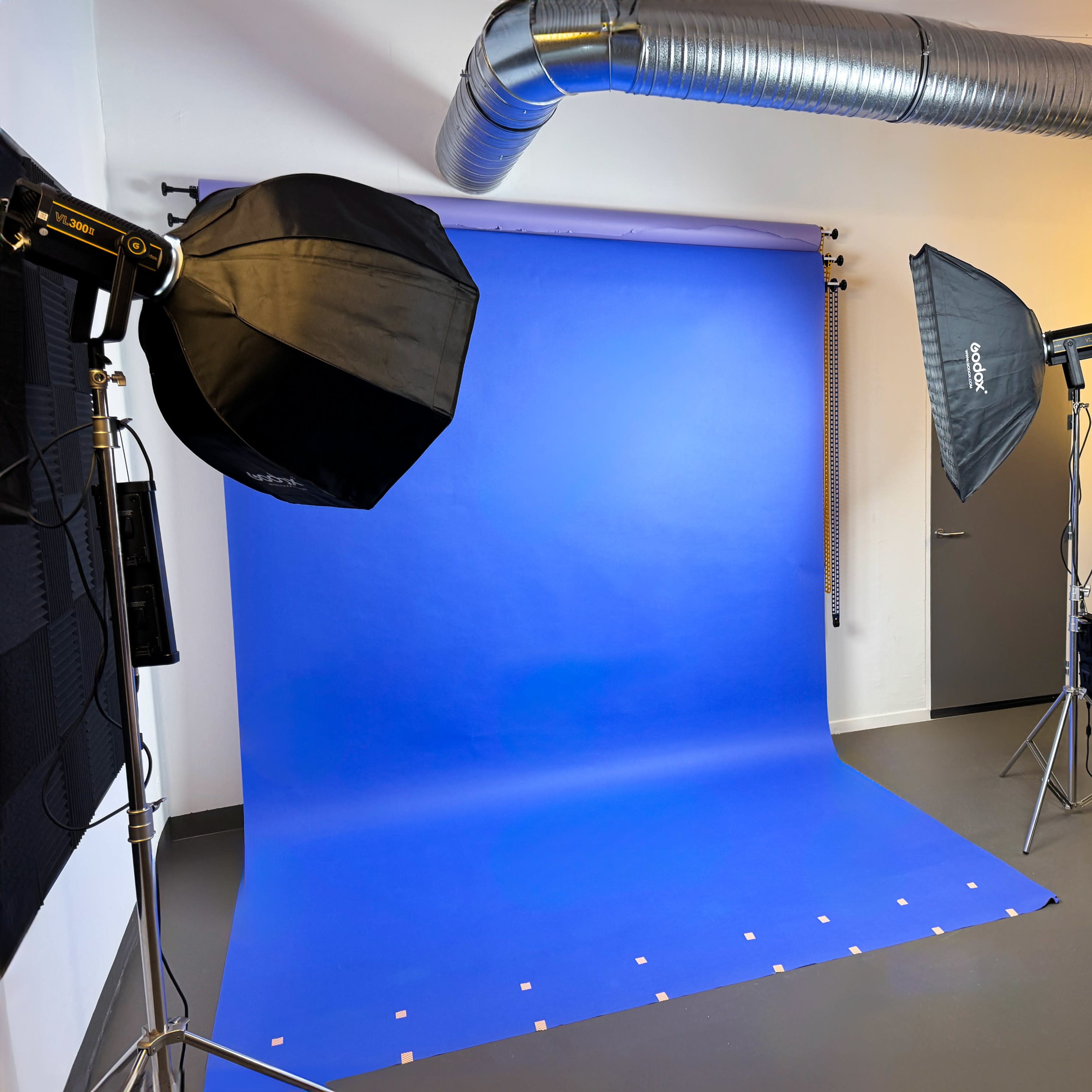 A photography studio with a blue backdrop, two softbox lights, and ventilation ducts visible on the ceiling.