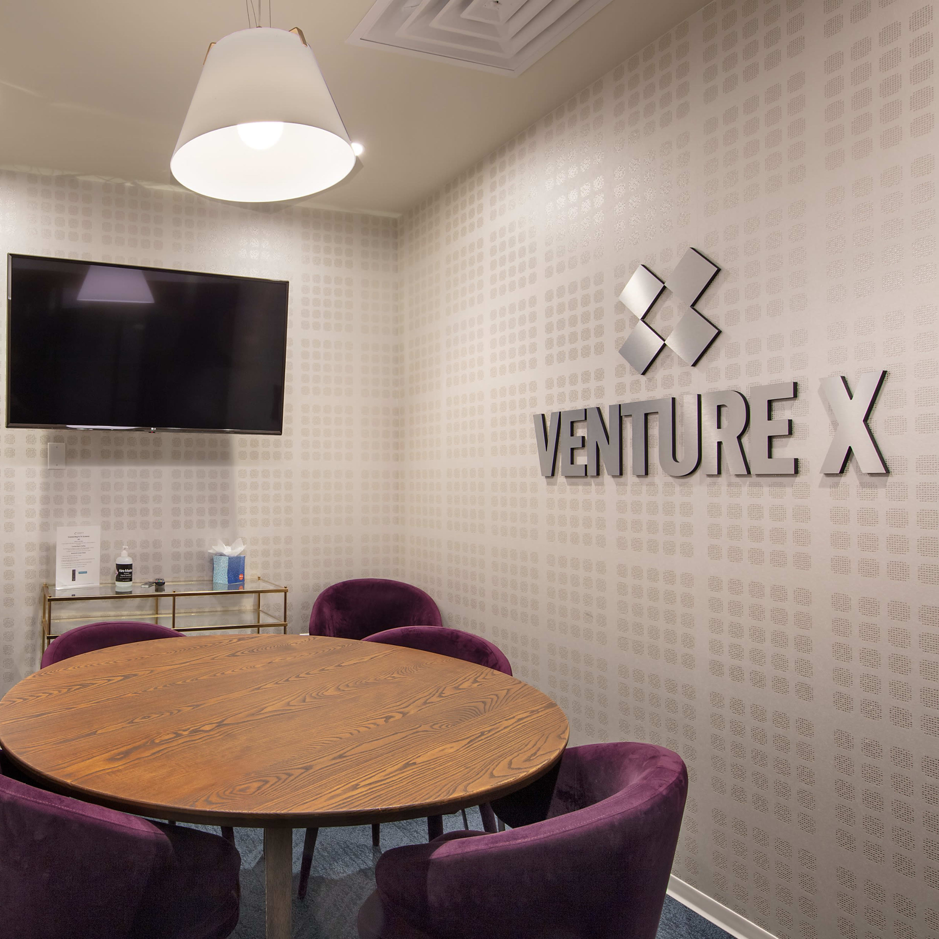A small conference room with a round wooden table, purple chairs, a wall-mounted television, and the "VENTURE X" logo displayed on the wall.
