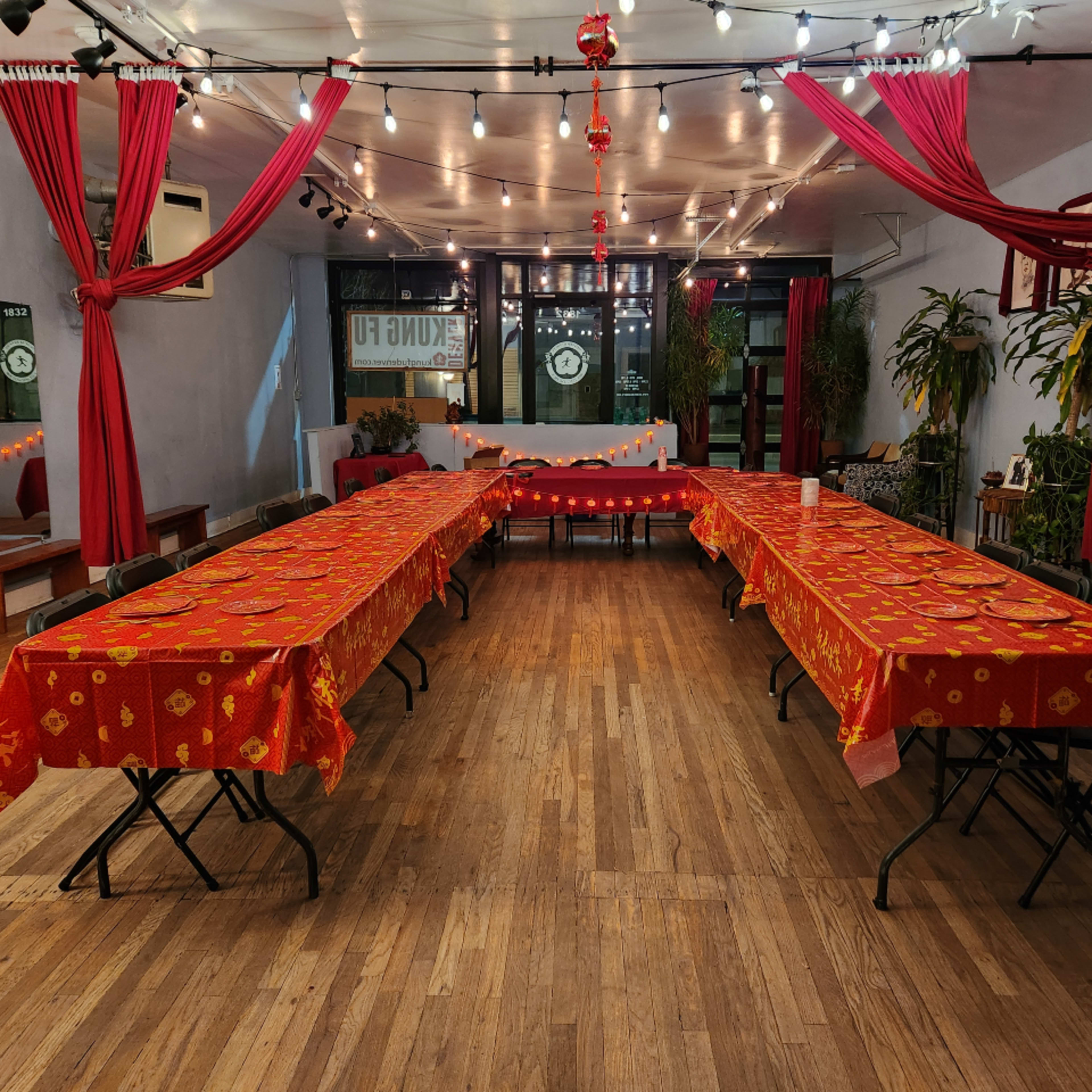 A spacious, decorated room with two long tables covered in red tablecloths, surrounded by vibrant curtains and string lights.