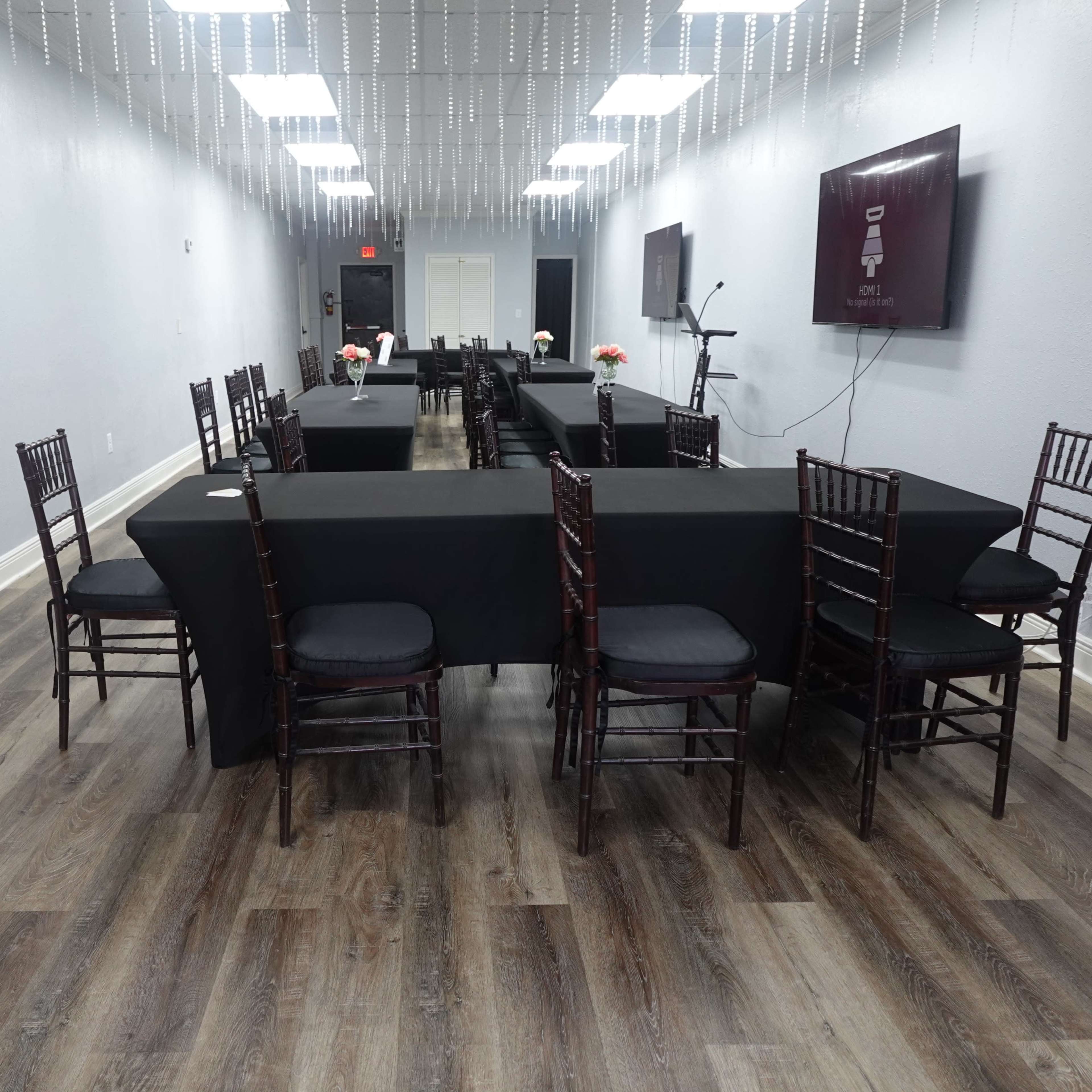 The interior of a spacious room features several rectangular tables covered with black tablecloths and surrounded by dark wooden chairs.
