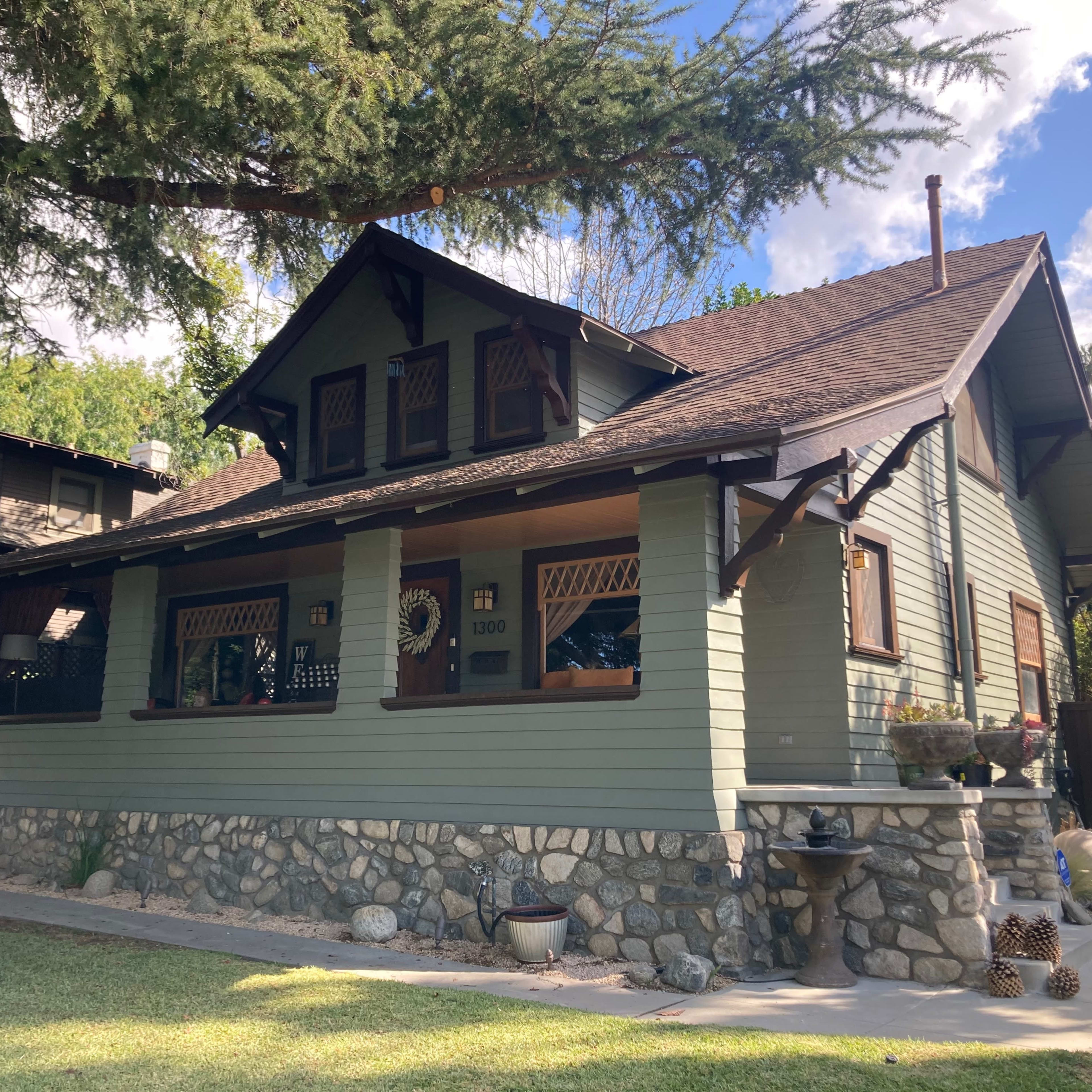 A two-story, green wooden house with a stone foundation and a front porch, surrounded by grassy landscaping and trees.