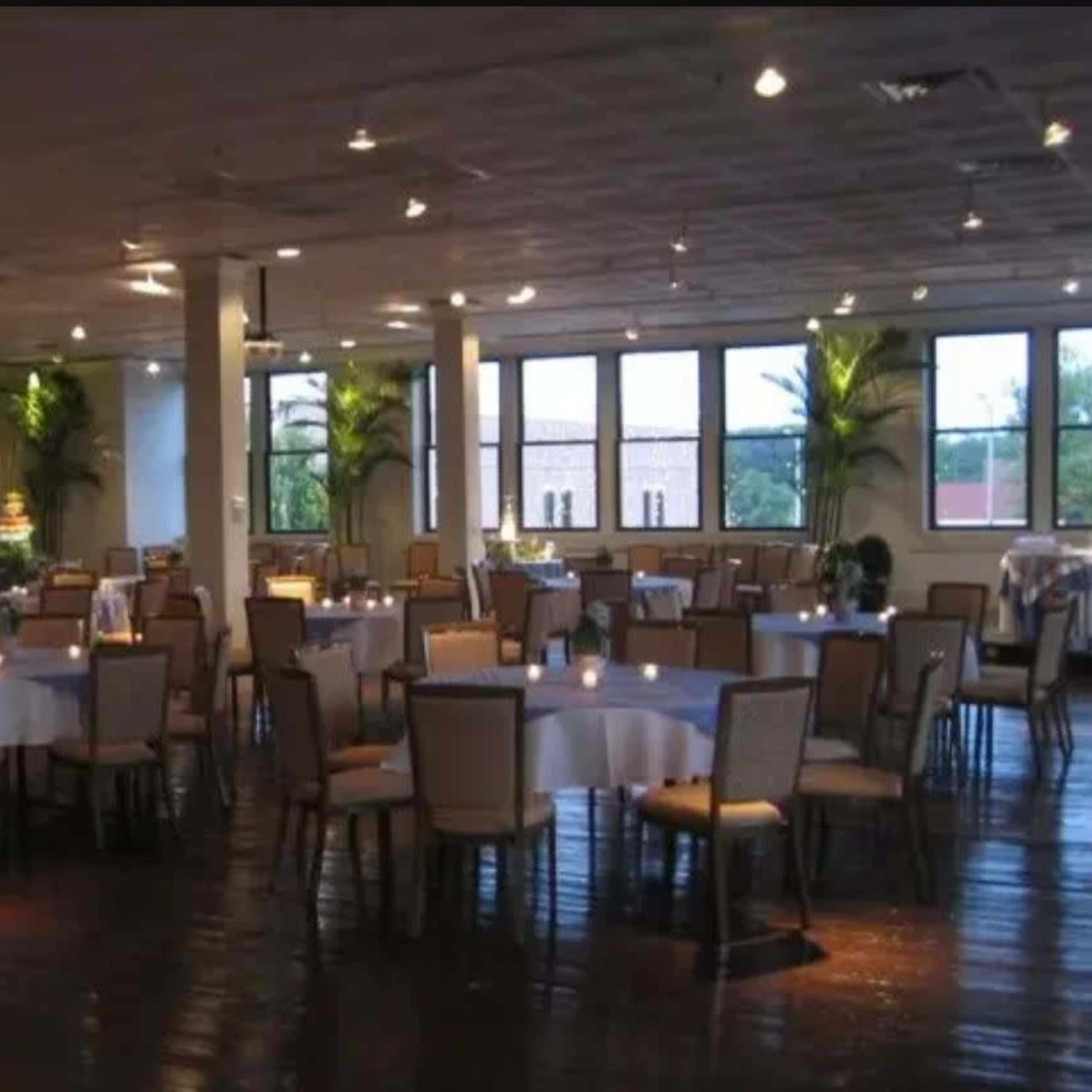 A spacious banquet hall with round tables set for dining, illuminated by warm lighting and featuring large windows with views of greenery outside.