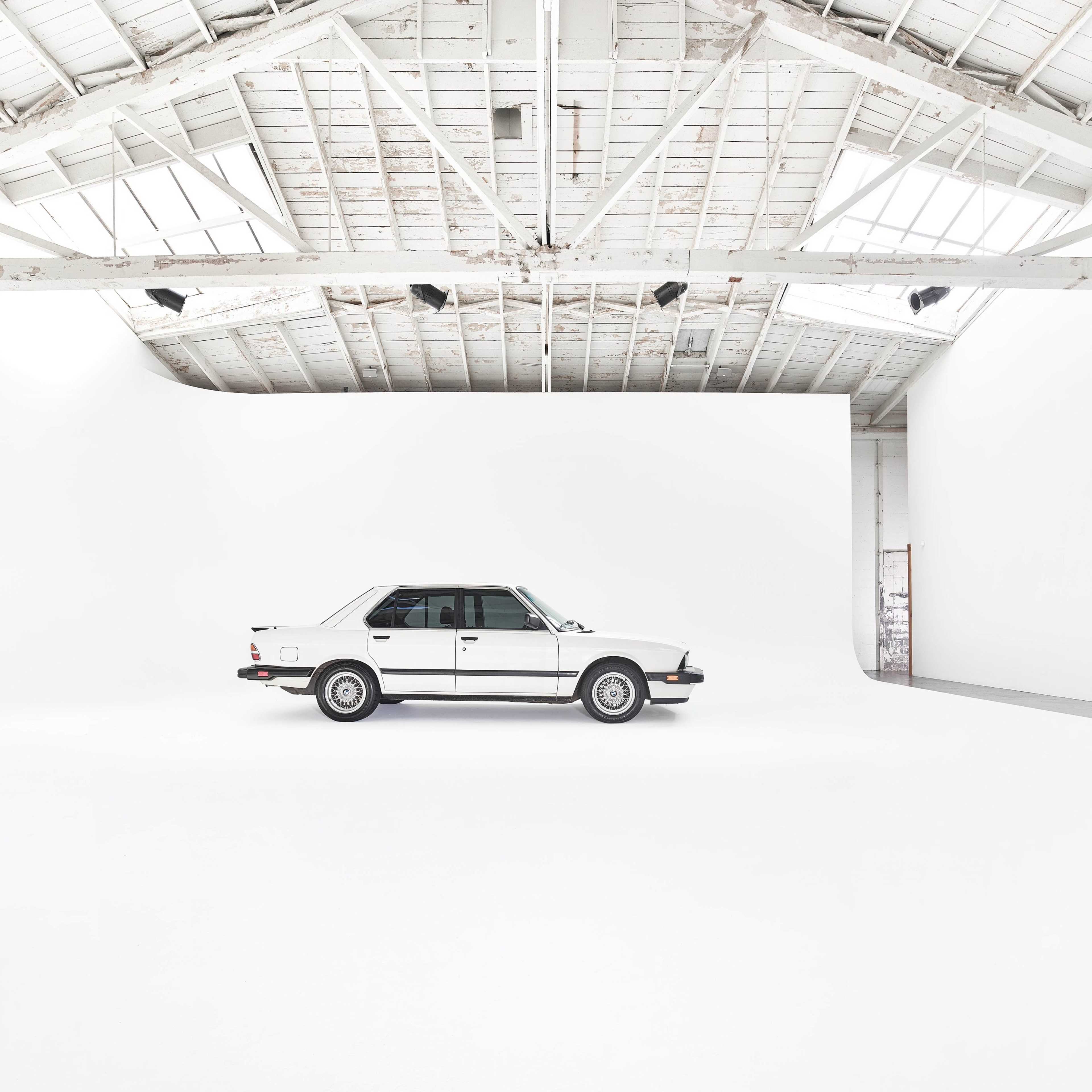 A white car is parked in a spacious, minimalist studio with a high ceiling and white walls.