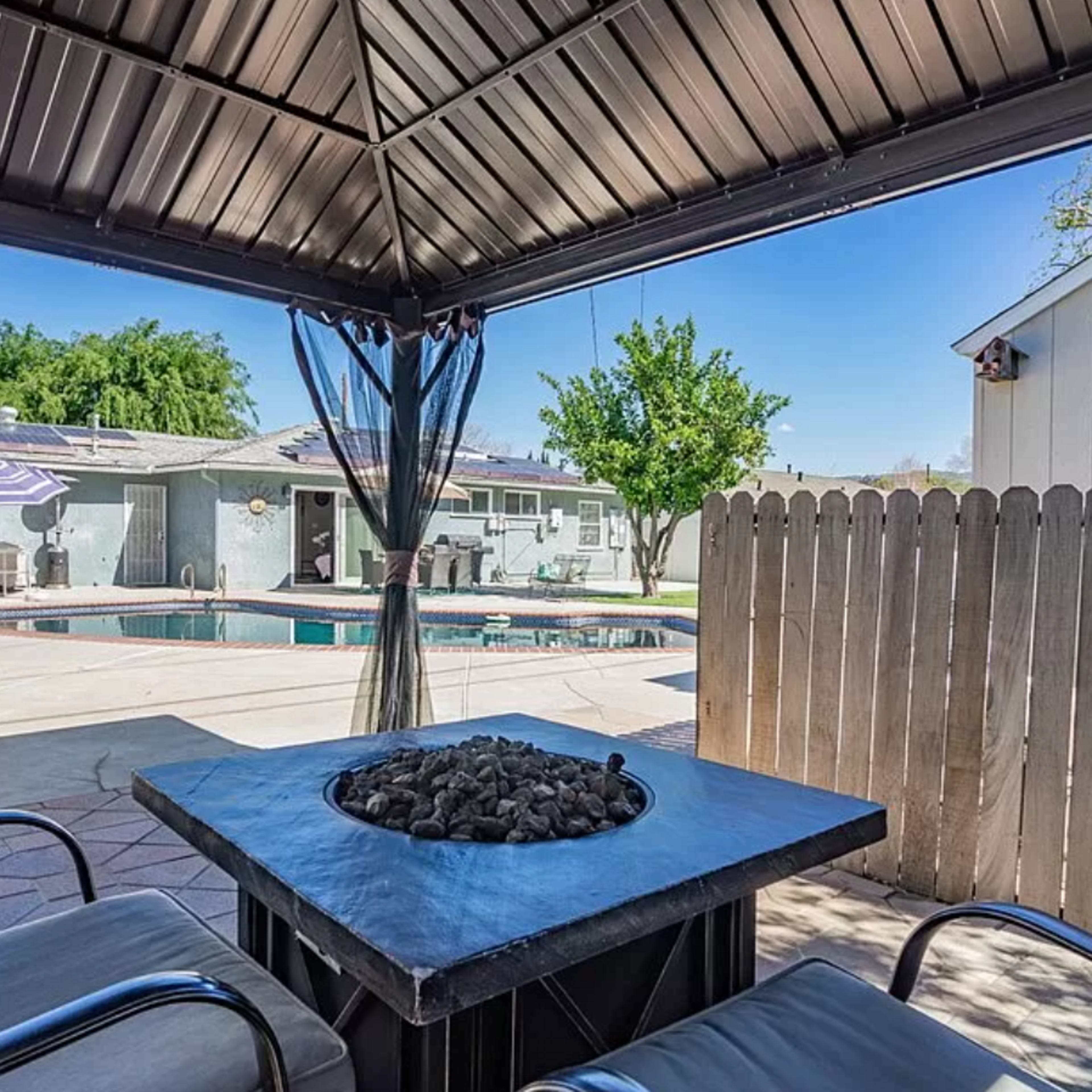 A backyard with a pool, a gazebo, and seating around a fire pit, enclosed by a wooden fence.