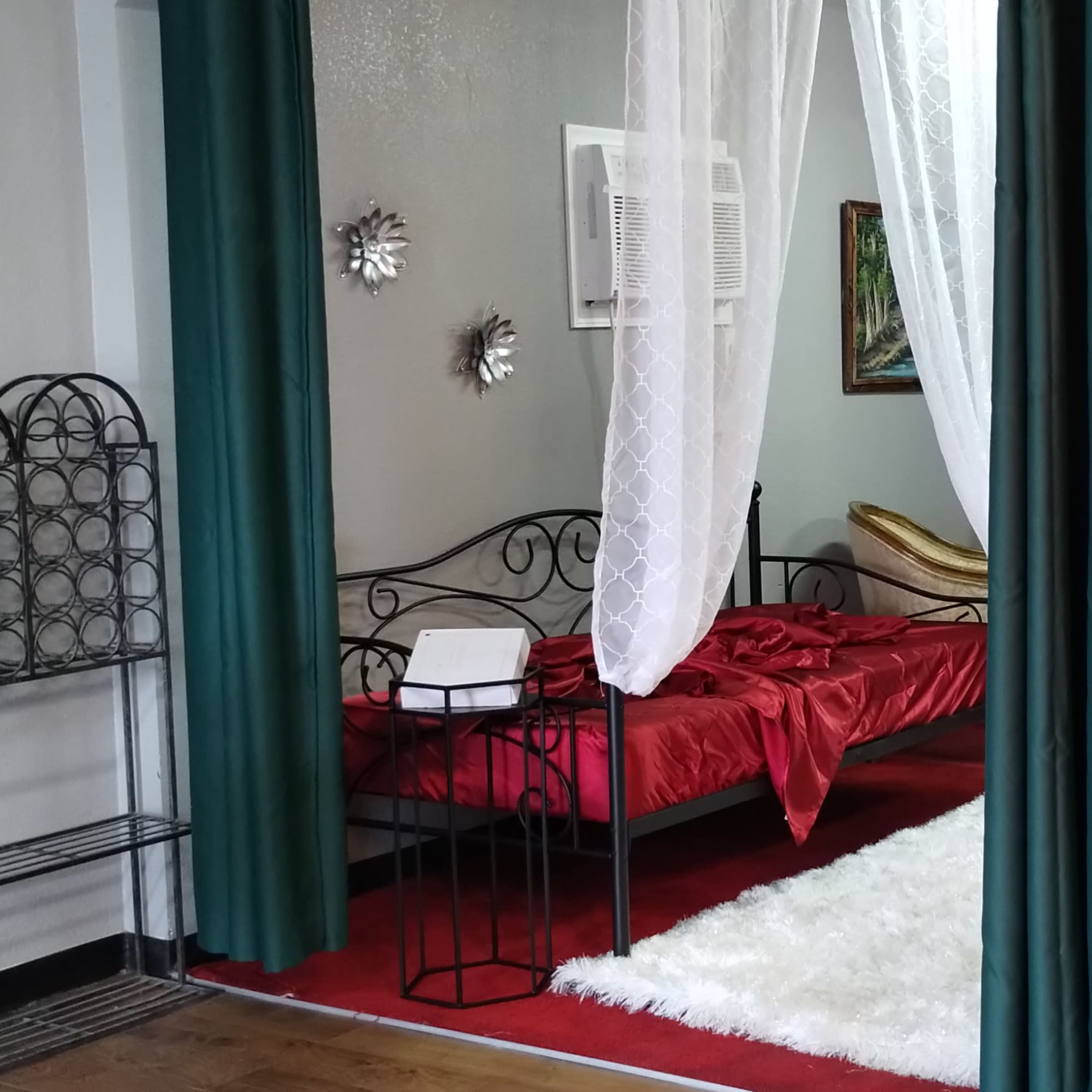 A room with a red carpet, a bed covered in red linens, white drapes, and a mix of furniture, including a chair and decorative wall art.