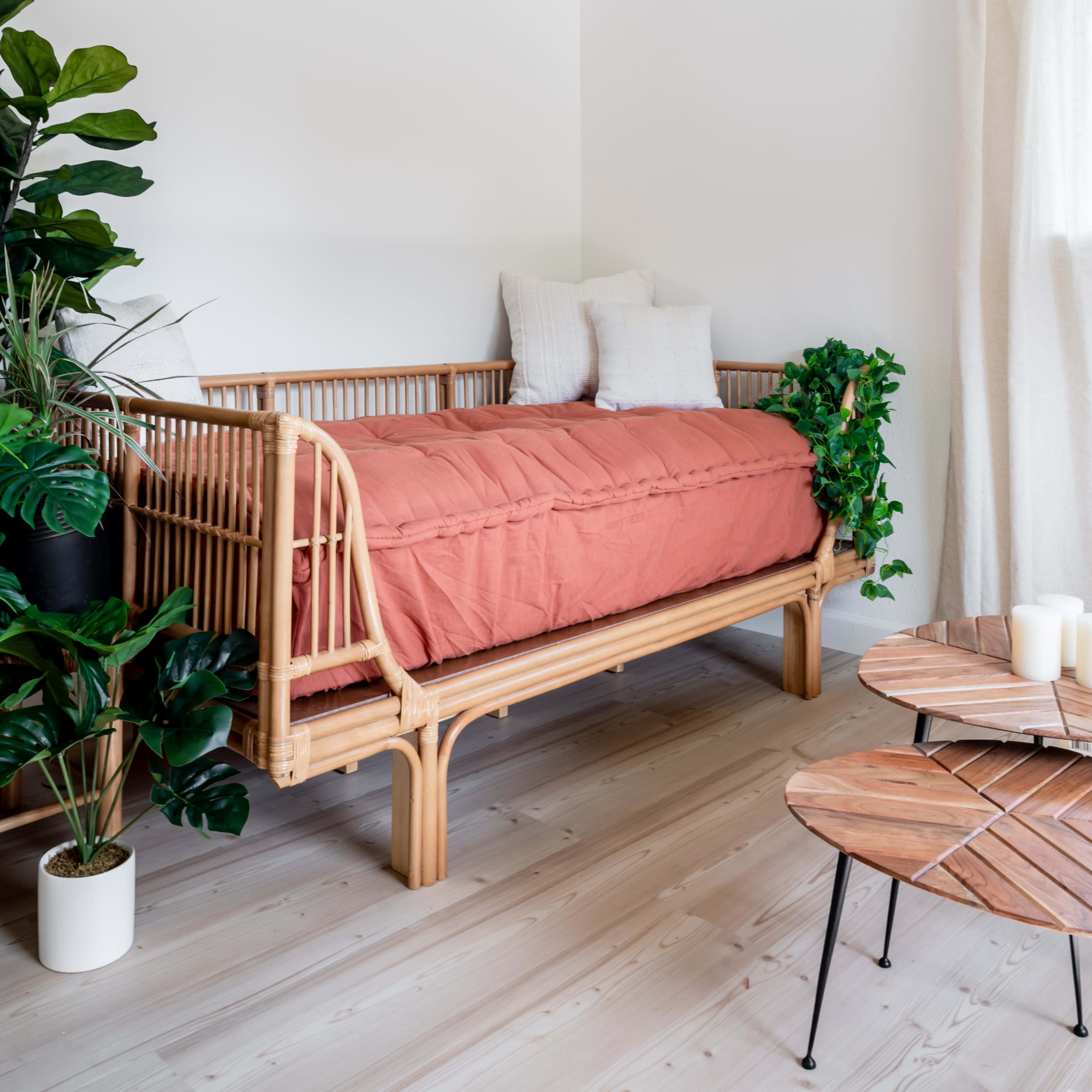 A room features a rattan daybed with a terracotta blanket, accompanied by potted plants and a wooden coffee table.