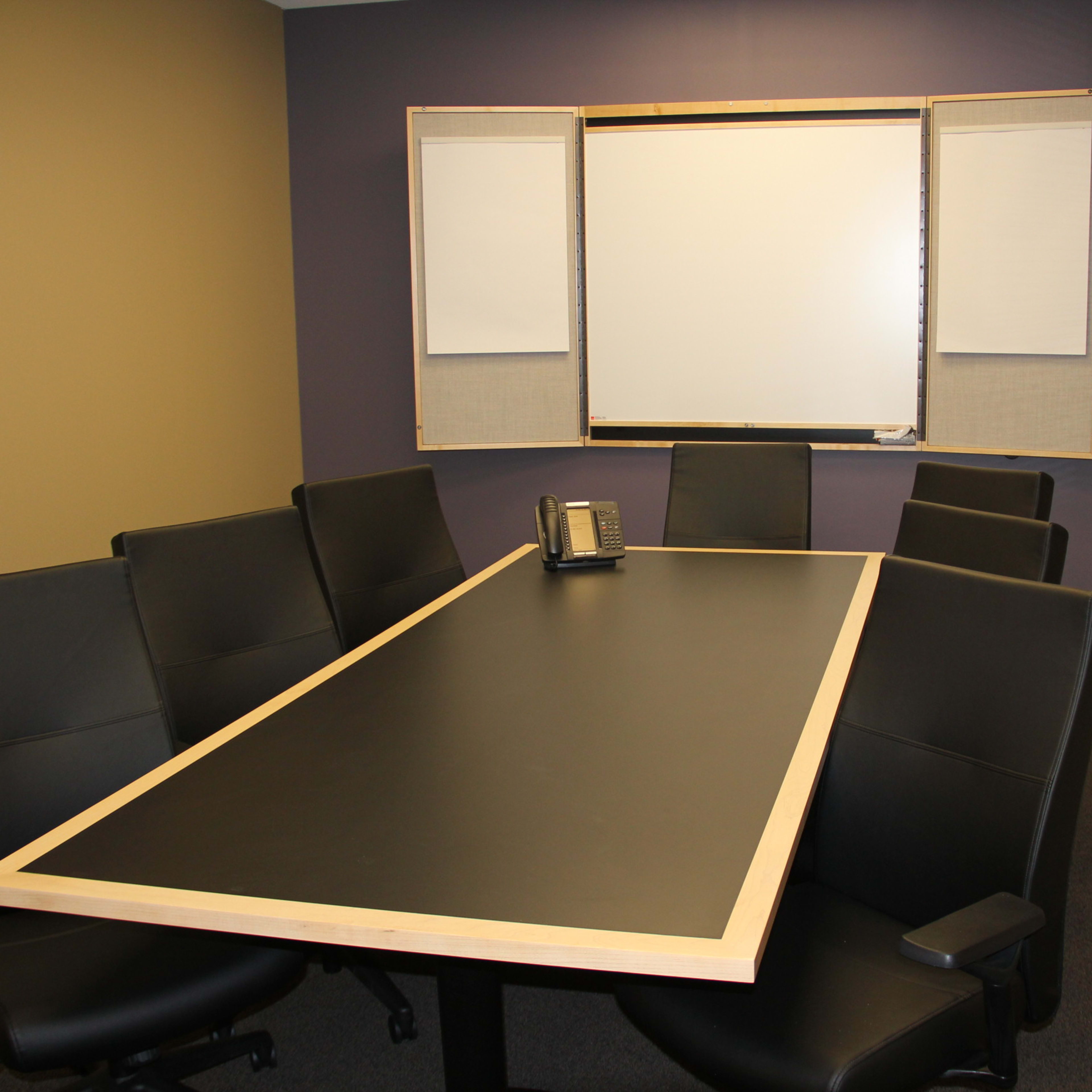A conference room features a large black table surrounded by eight black chairs, with a whiteboard and door visible in the background.