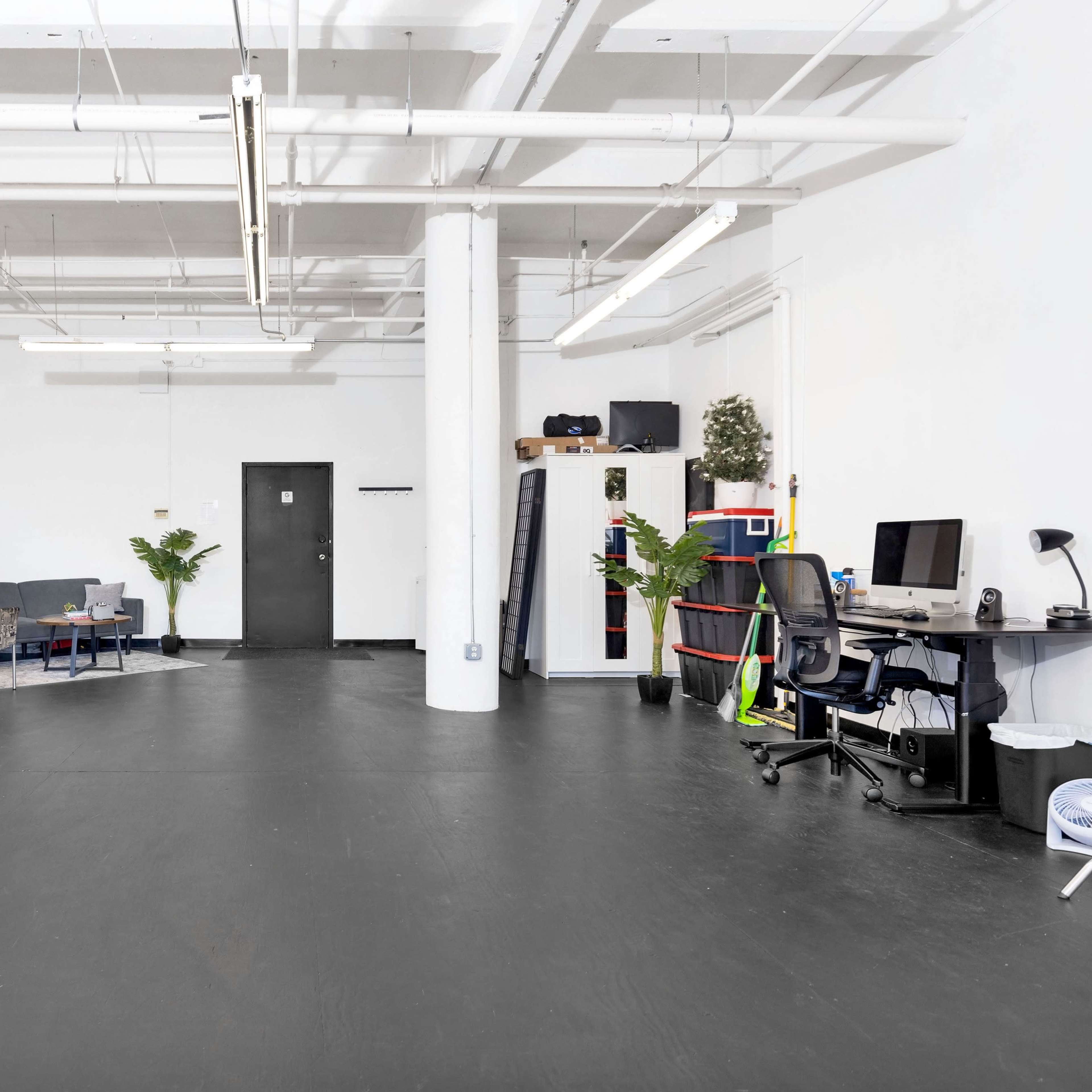 A spacious, modern room with black flooring, featuring seating areas, plants, a desk, and clothing racks along the walls.