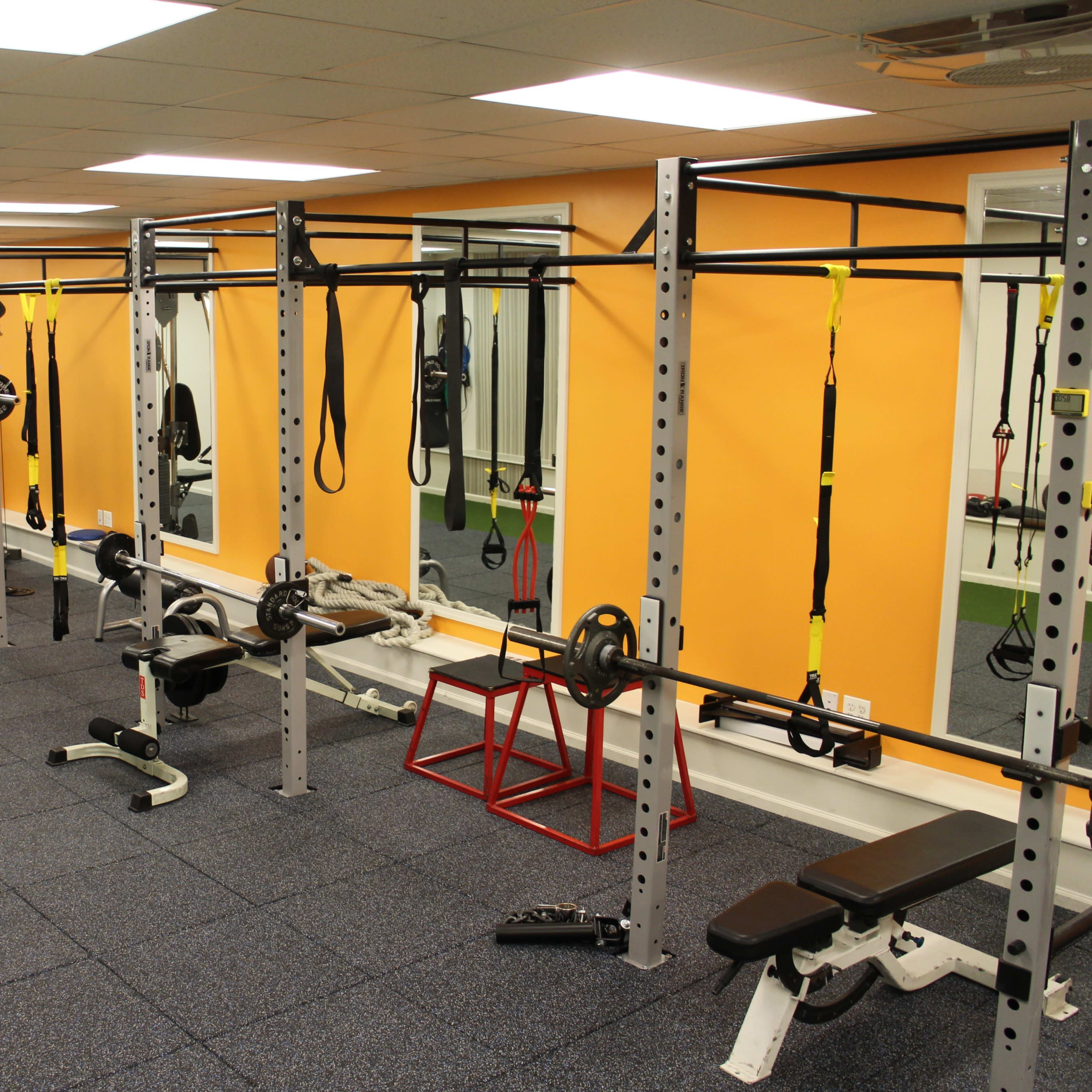 The gym features various weightlifting equipment and a functional training area against bright orange walls.
