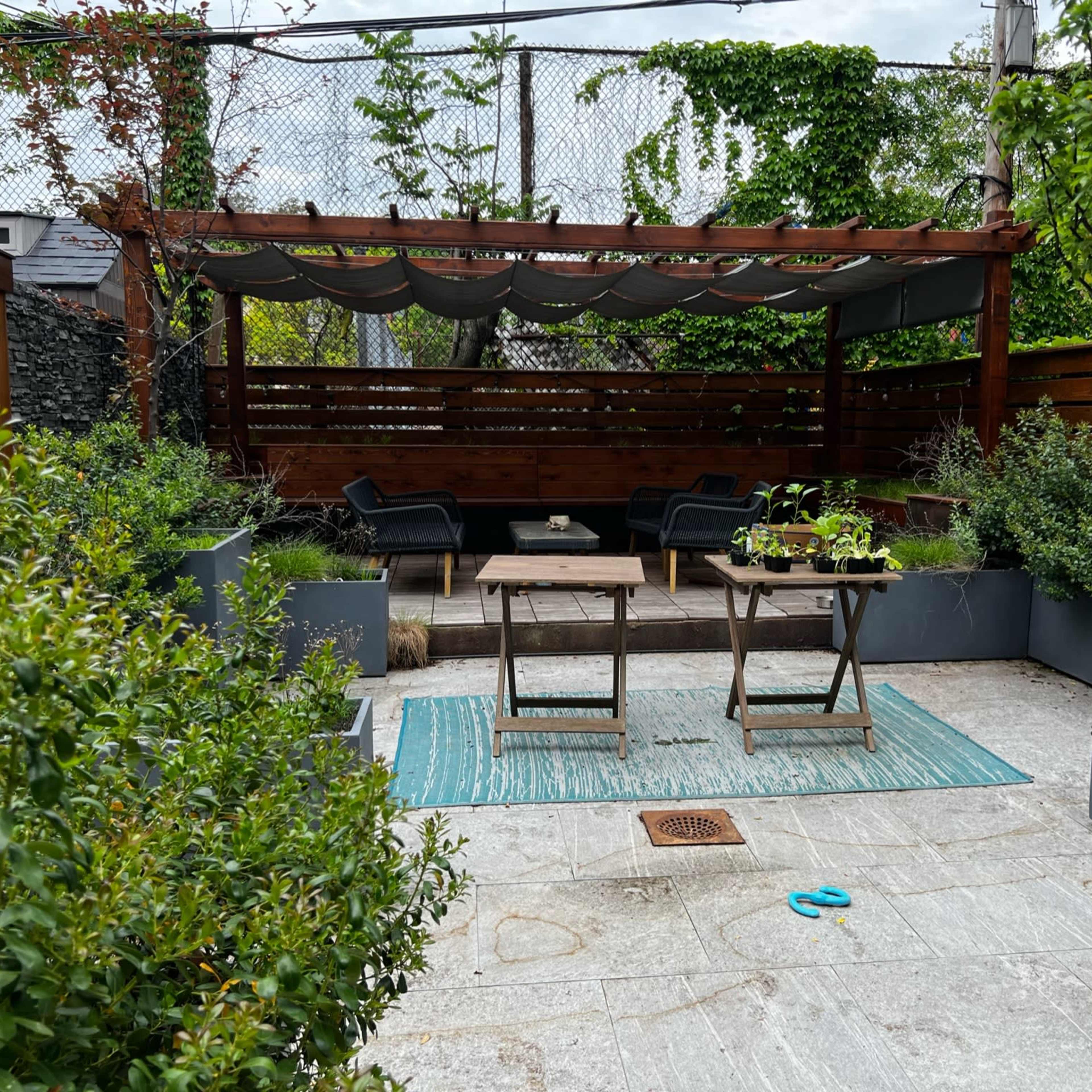 A small outdoor patio area featuring a wooden pergola, two tables, and several potted plants arranged around a stone pathway.