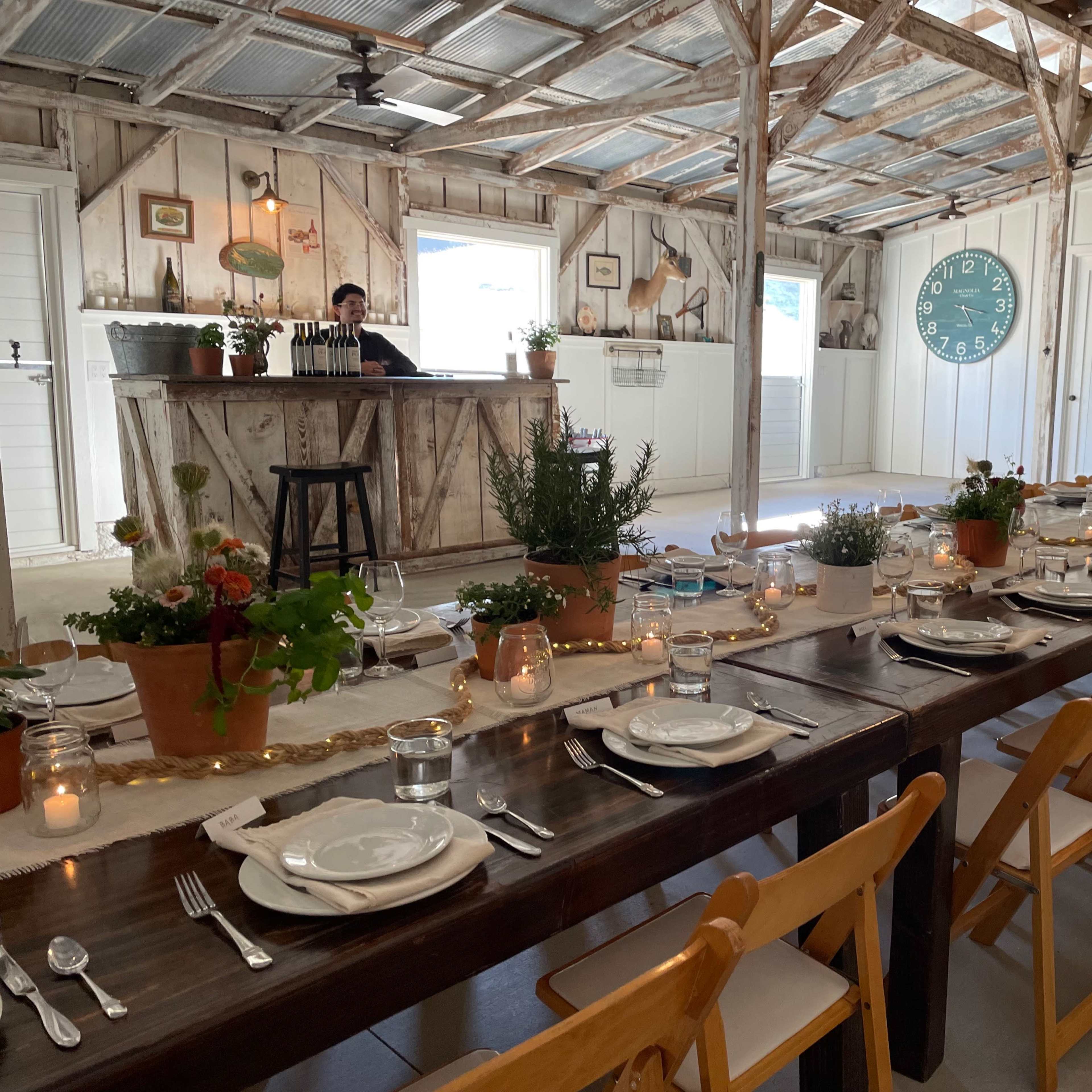 A long wooden table is set for a meal in a rustic indoor space featuring a bar, potted plants, and a large clock on the wall.