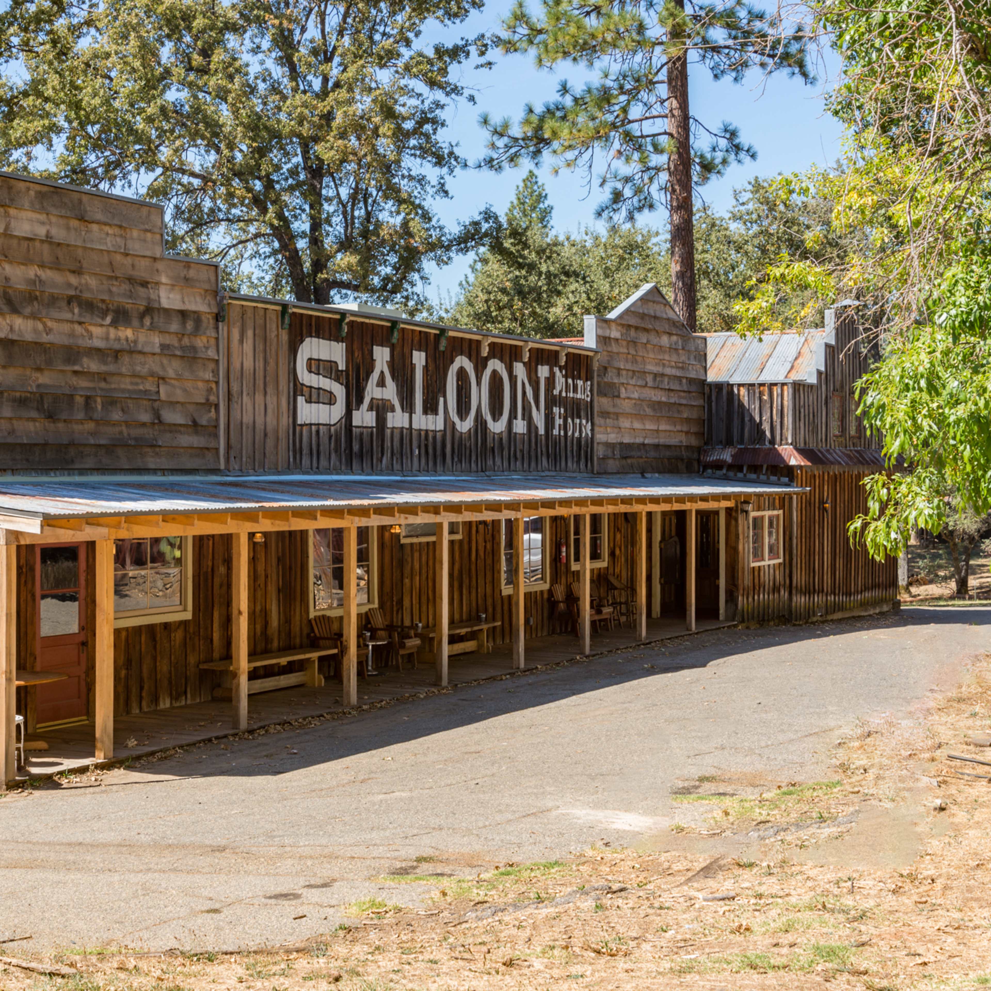 A wooden saloon building with a covered porch is located along a winding path surrounded by trees.