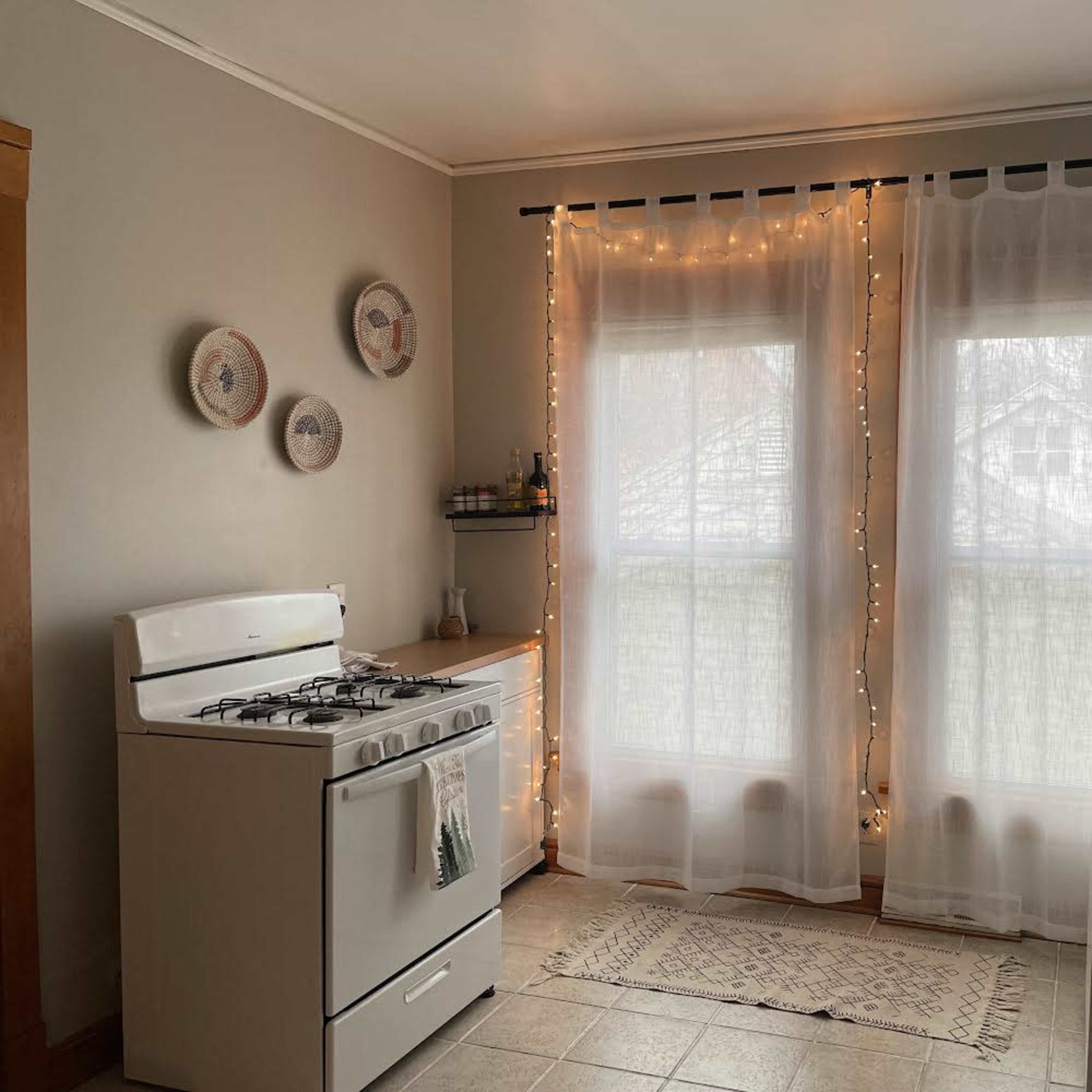 A kitchen with a white stove, a refrigerator, and large windows dressed with sheer curtains and string lights.