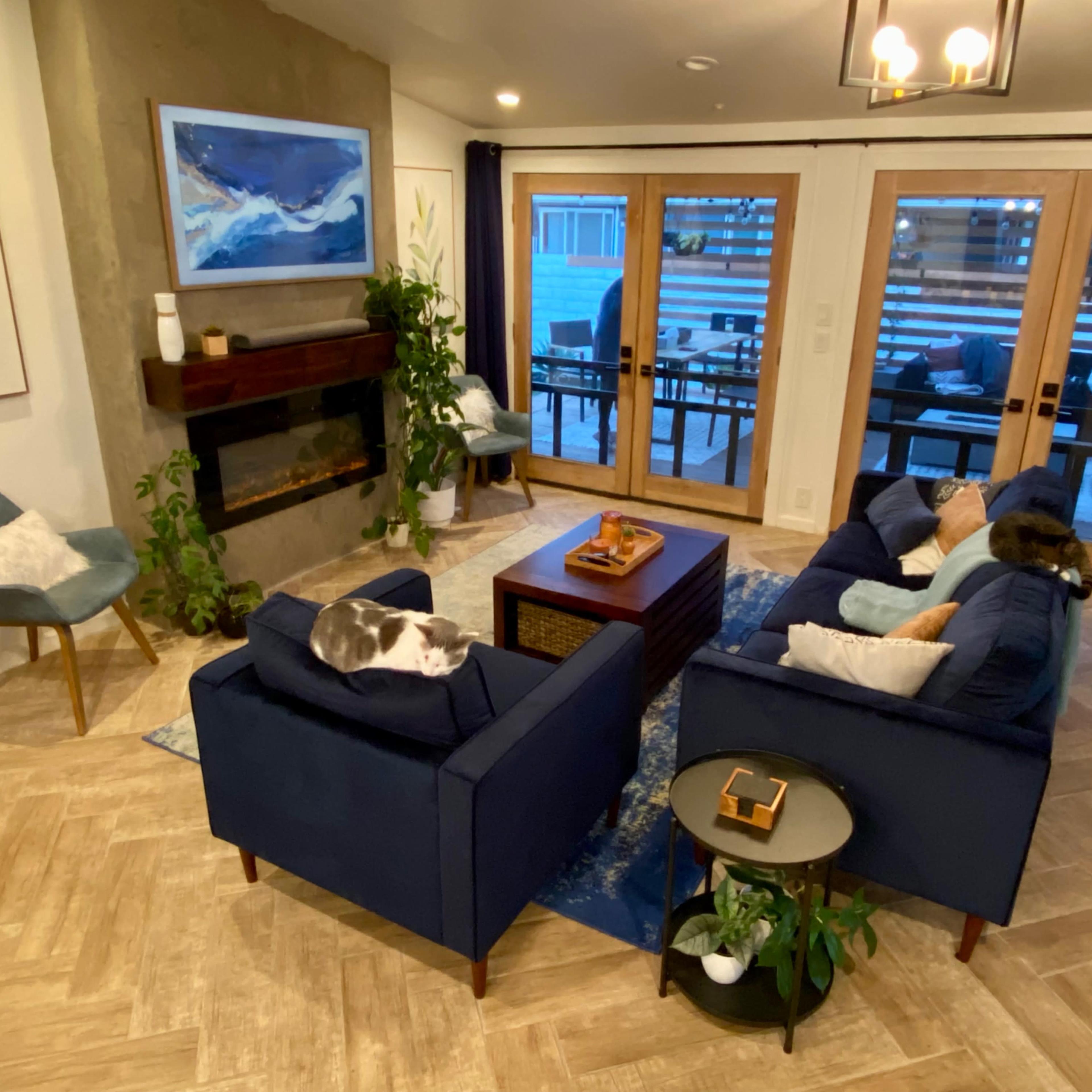 A living room with two blue sofas facing each other, a coffee table in between, a fireplace on the wall, and large glass doors leading to an outdoor area.