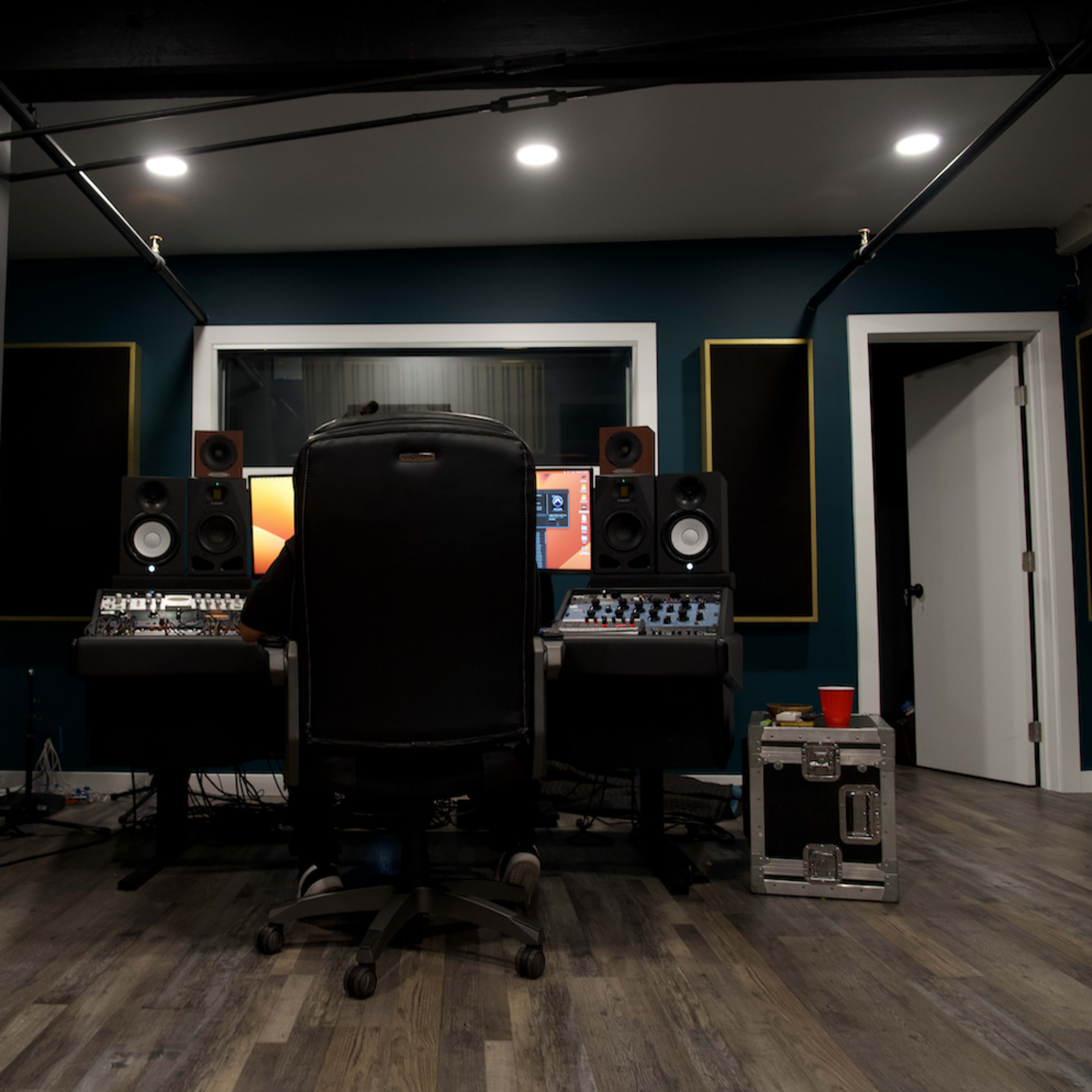A person sits at a recording studio desk with audio equipment and speakers, facing a computer screen in a dimly lit room.