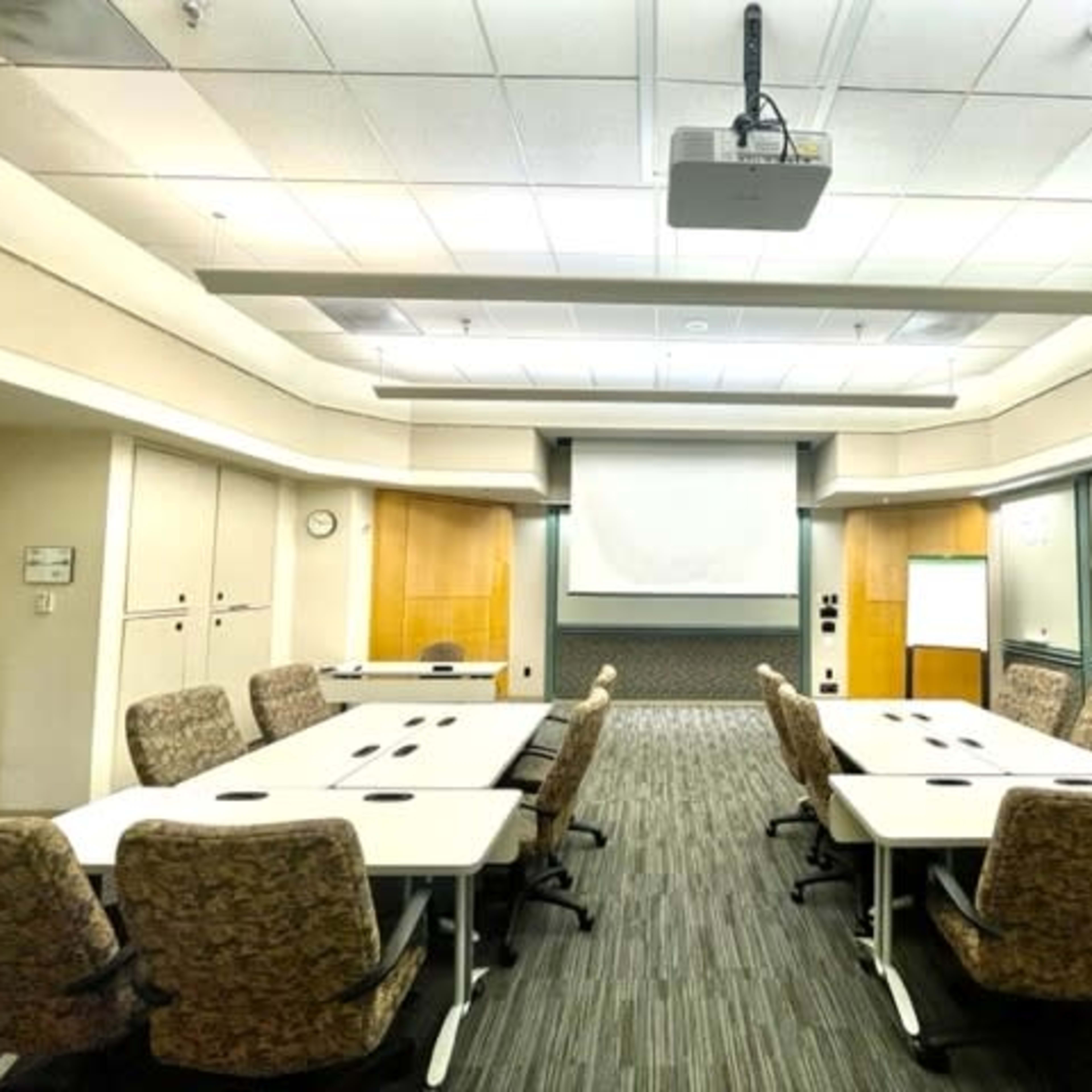 A conference room is set up with several tables arranged in a U-shape and chairs along the sides, featuring a projector and a large screen at the front.