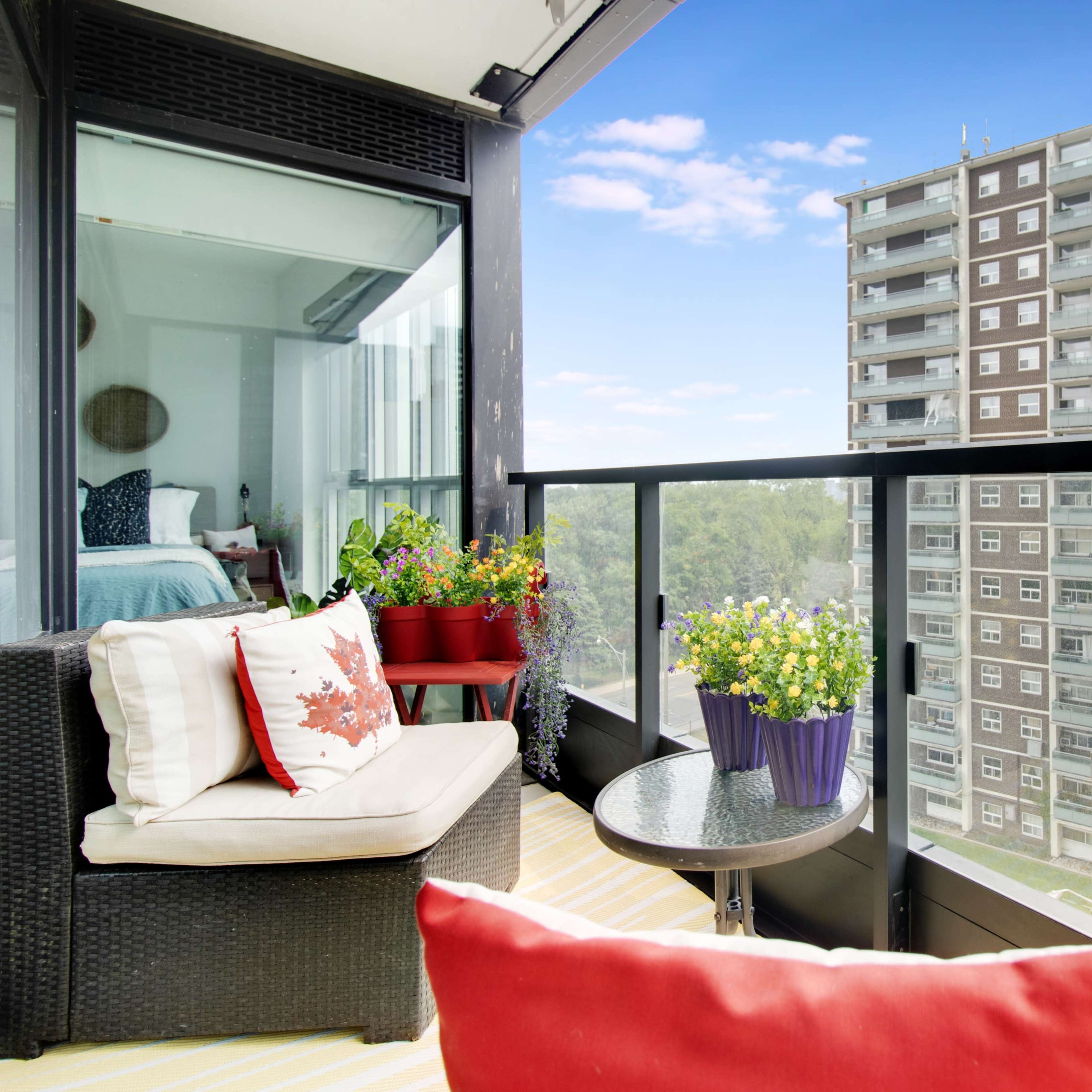 A balcony with potted plants and outdoor furniture overlooks an apartment building and a clear blue sky.