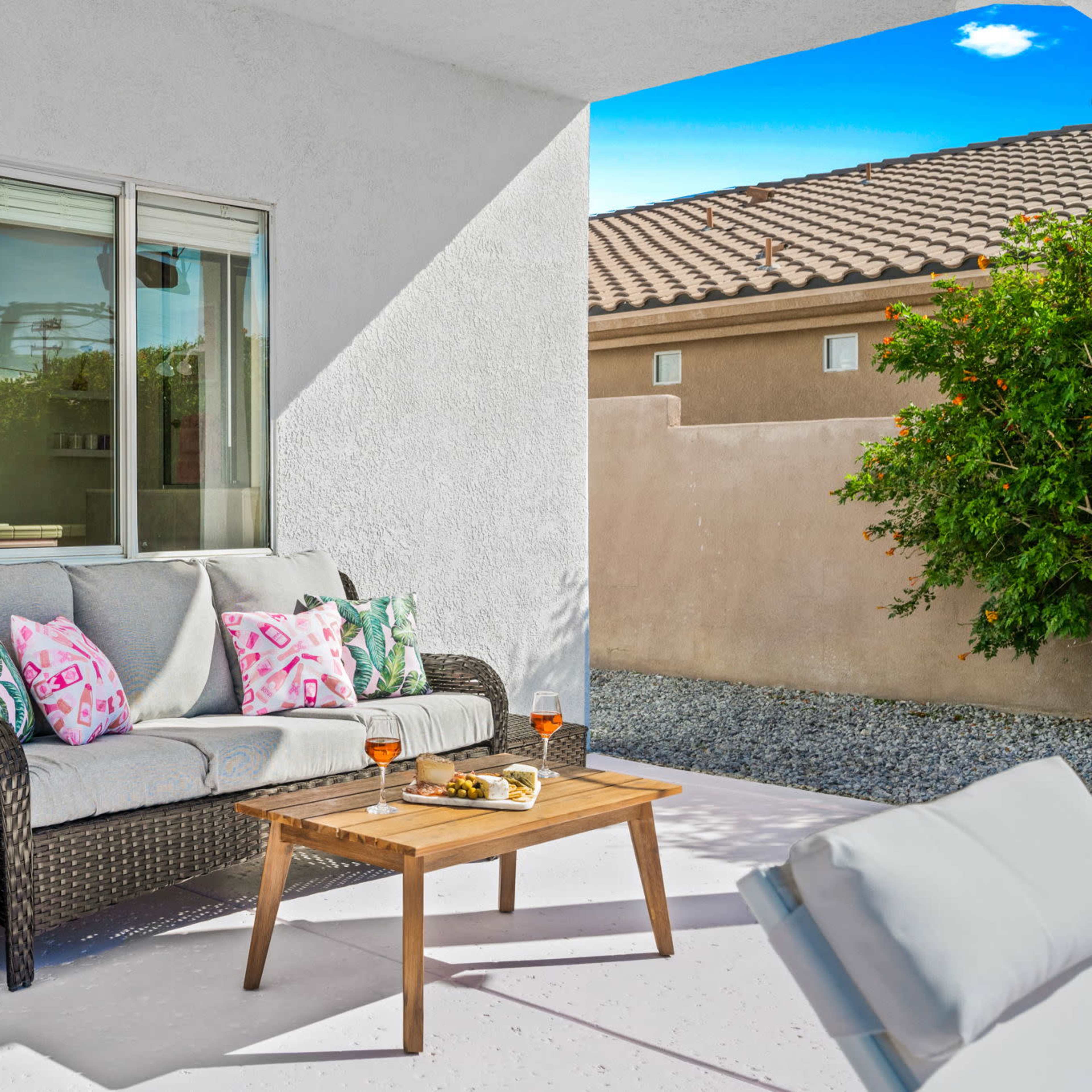 A patio area features a wicker sofa with decorative pillows and a wooden coffee table set with snacks and drinks, surrounded by a stone landscape and a house wall.