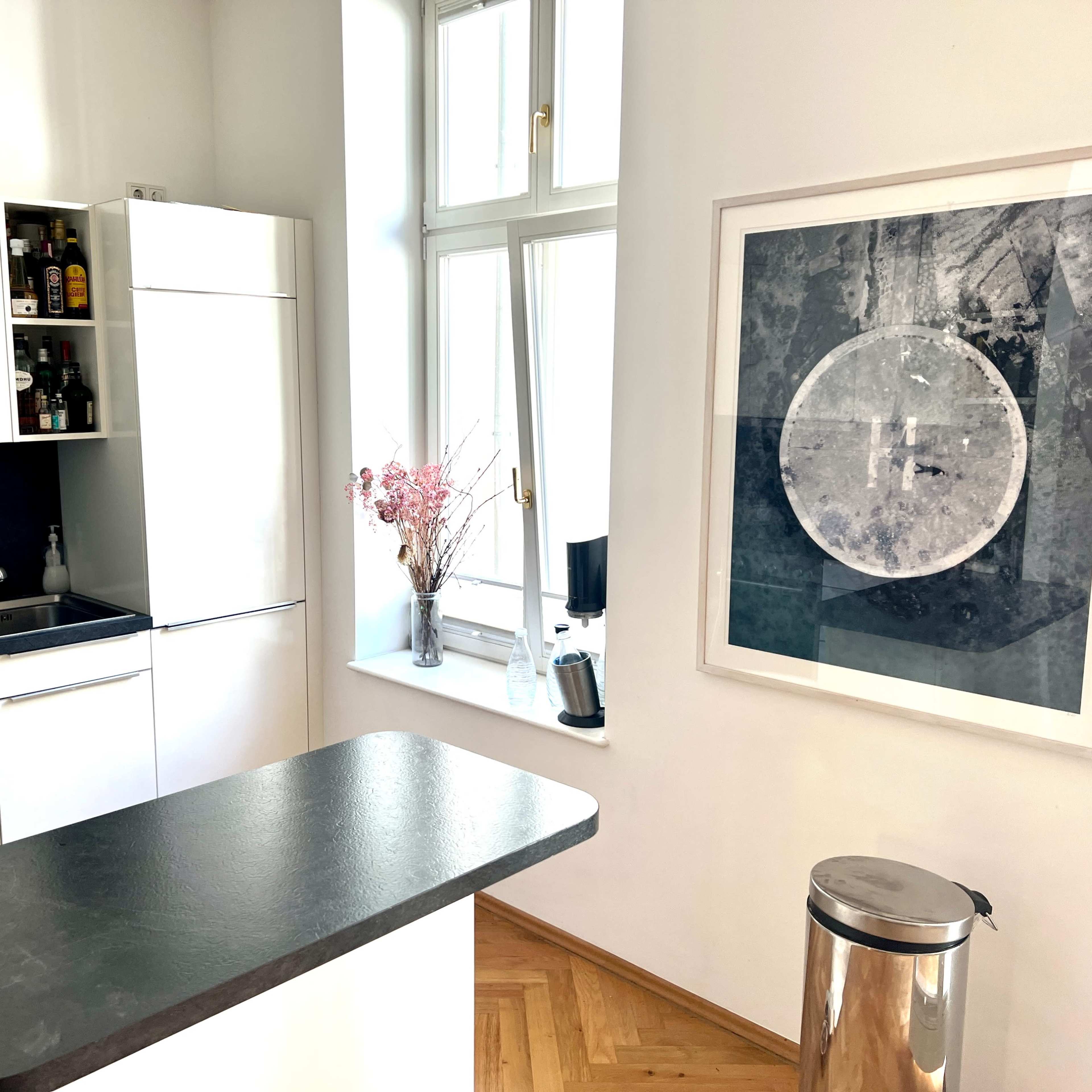 A modern kitchen with white cabinetry, a black countertop, and a large framed artwork on the wall beside a window.