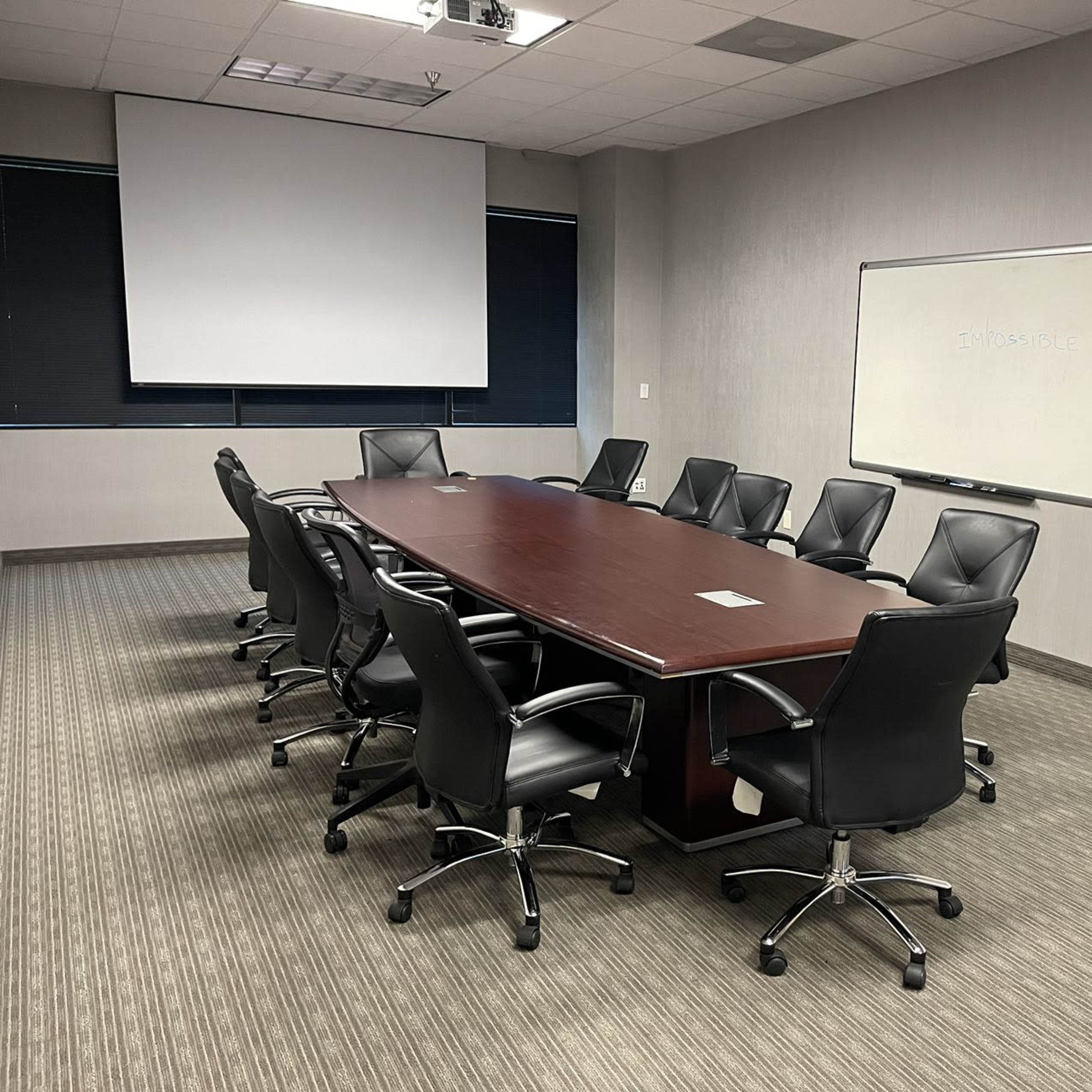 A large, unoccupied conference room features a long wooden table surrounded by black swivel chairs, with a projector screen and whiteboard in the background.