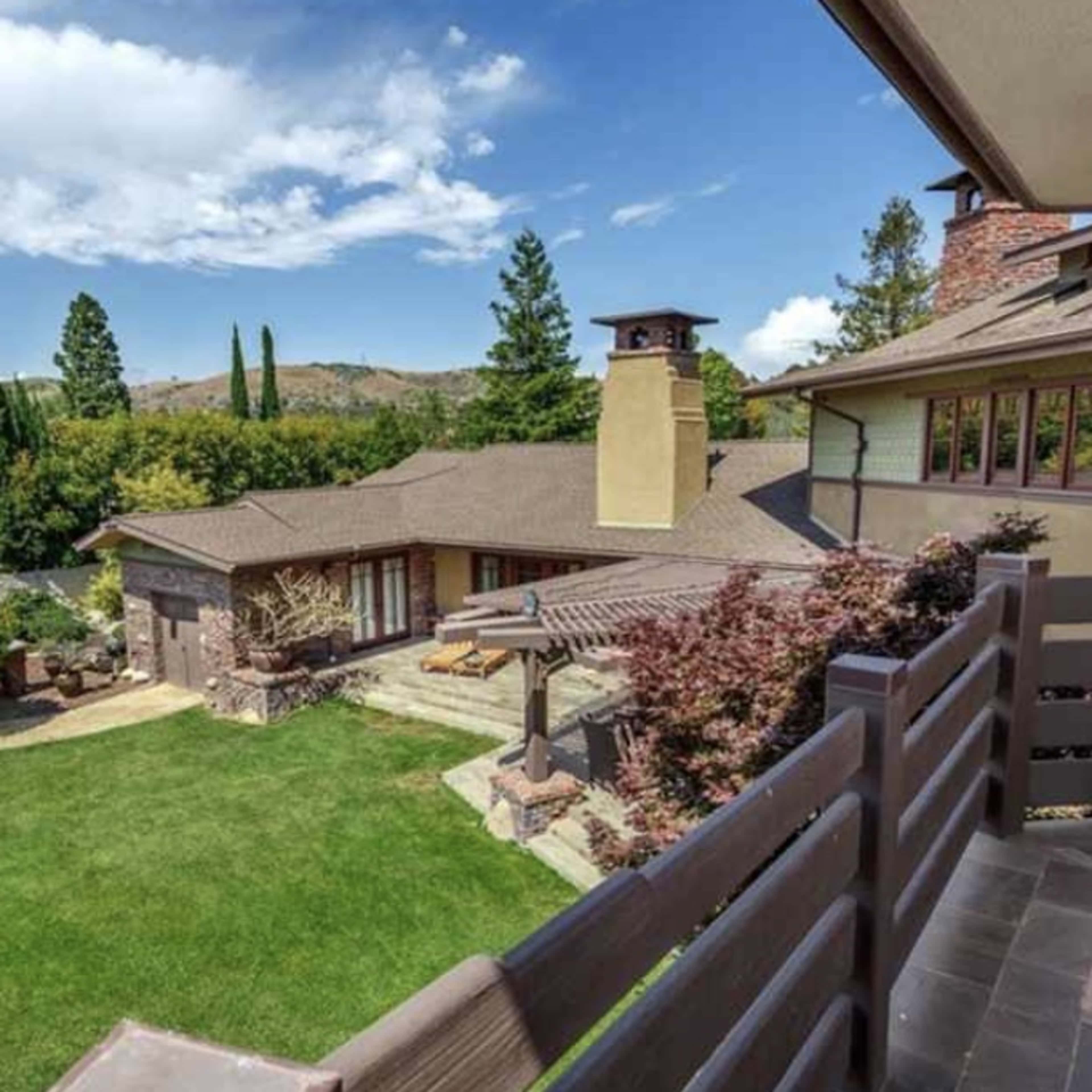 A modern house with a landscaped yard and patio, viewed from a balcony against a backdrop of blue skies and trees.