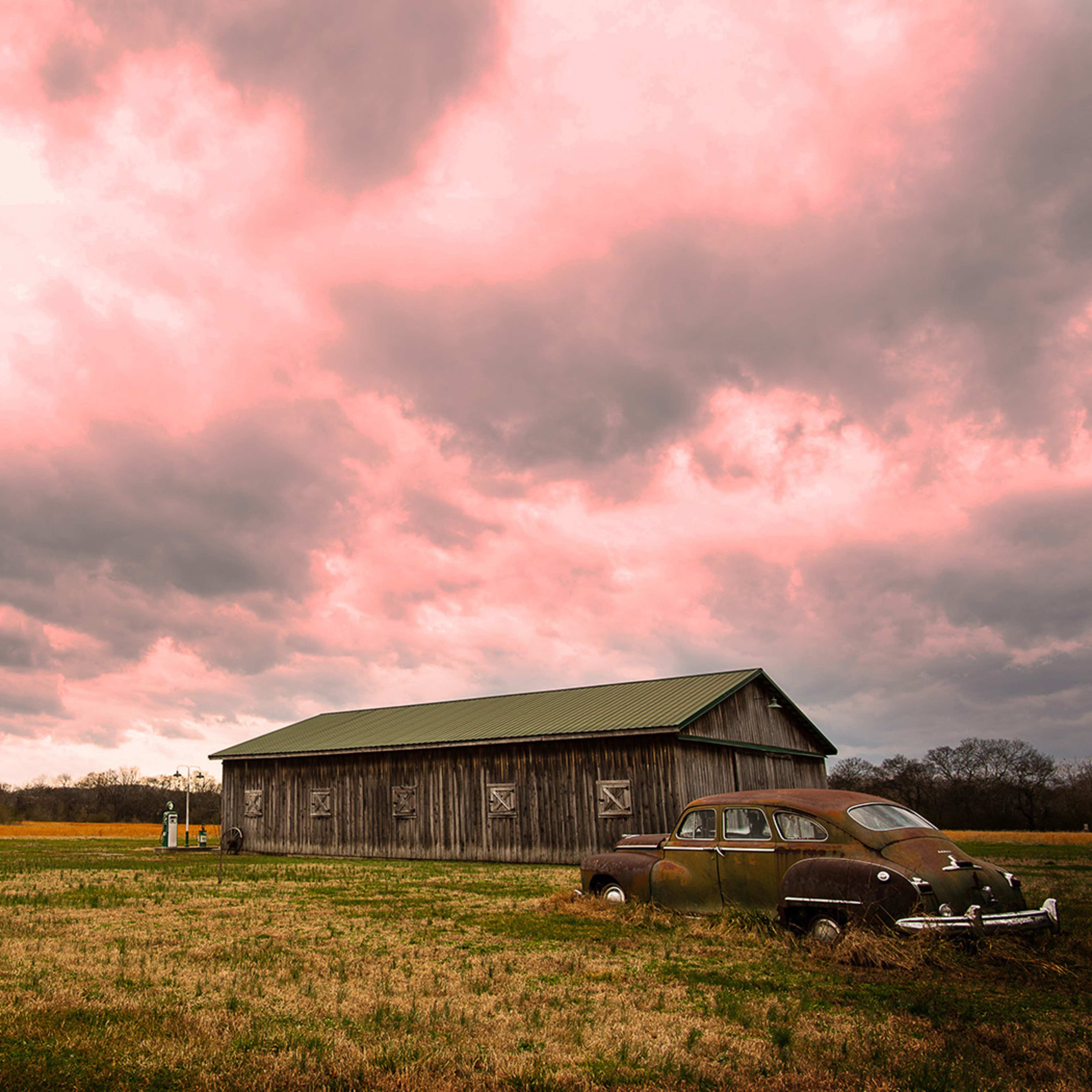 A weathered barn stands beside an old car in a grassy field under a dramatic pink and gray sky.