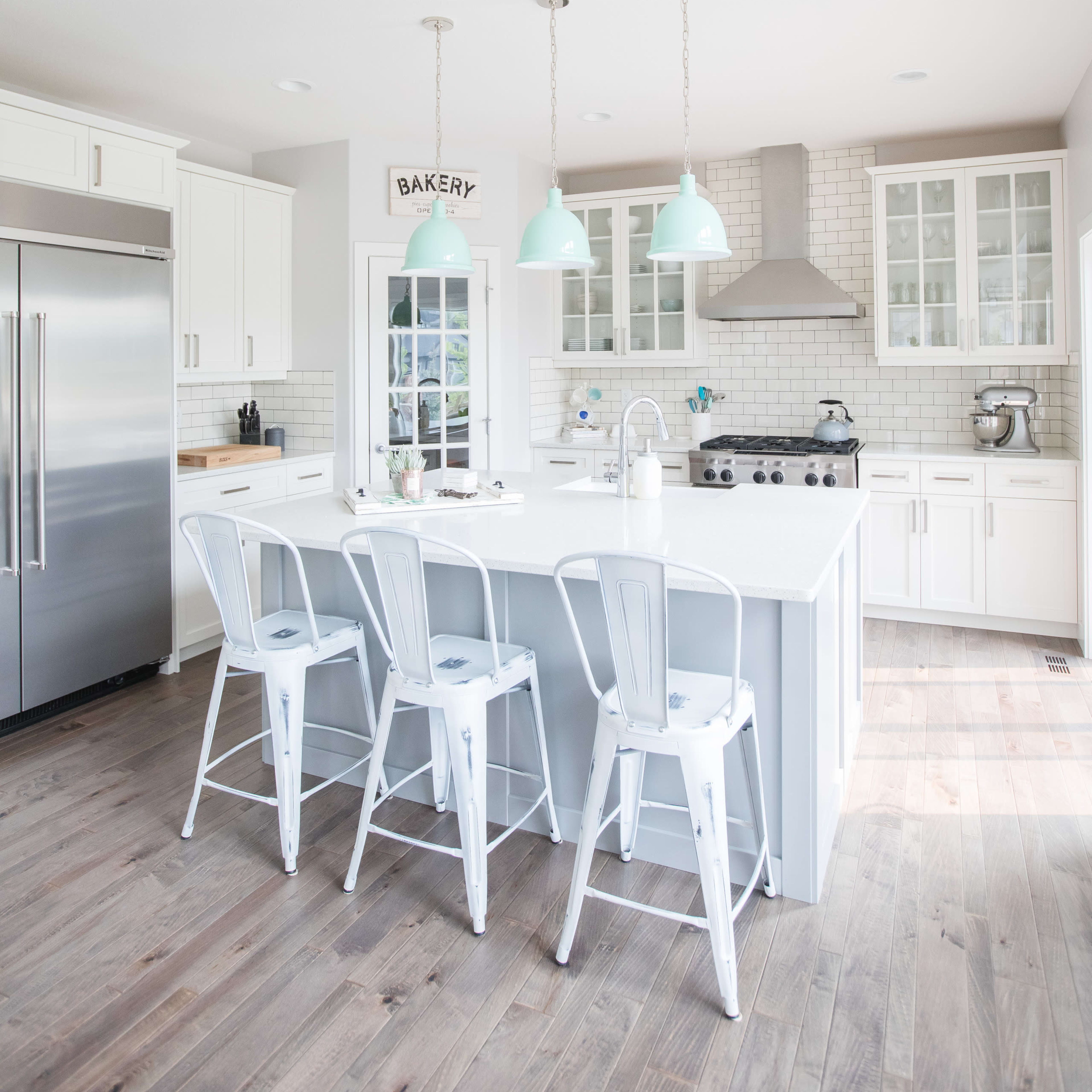A modern kitchen features white cabinets, stainless steel appliances, a large island with seating, and pendant lights hanging above.