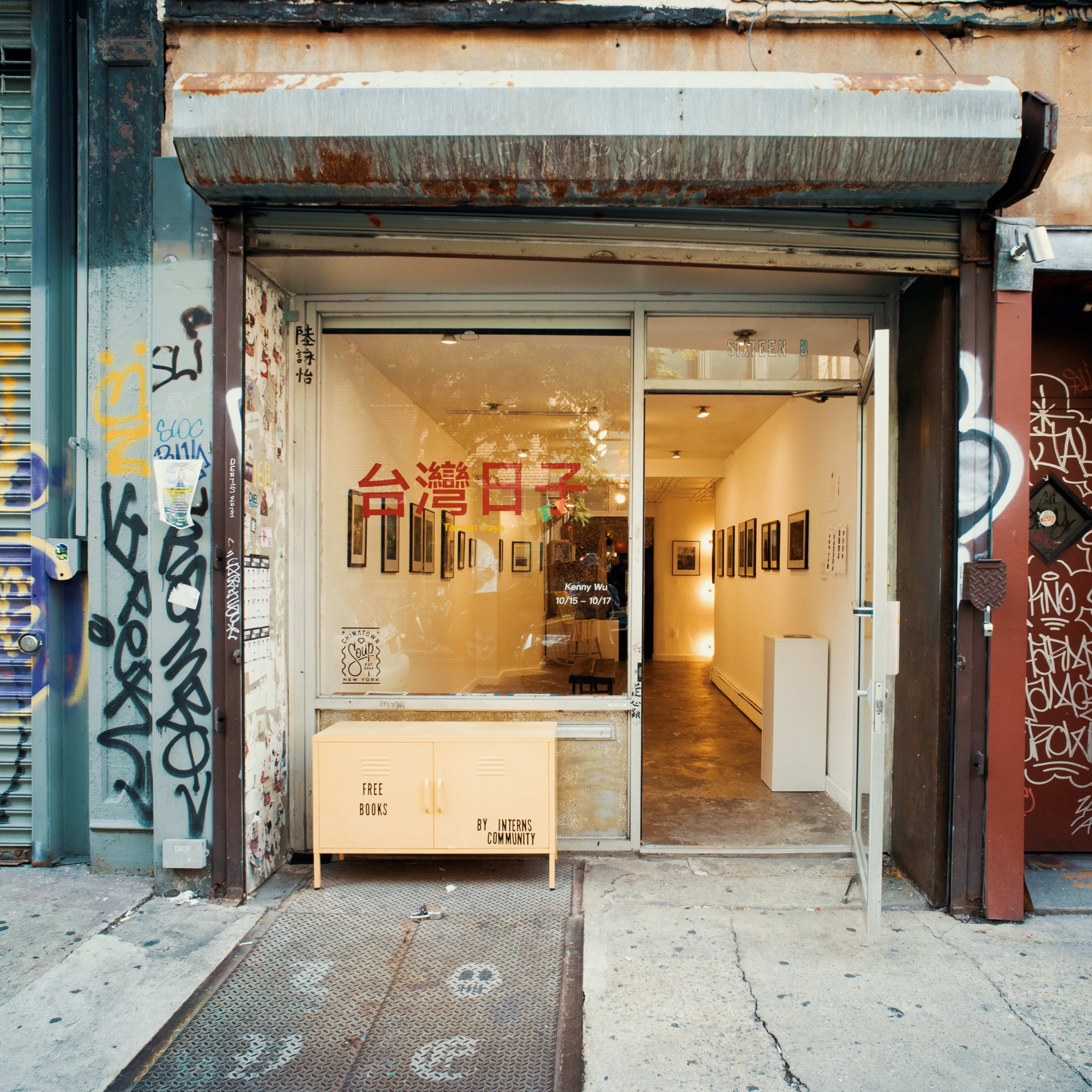 A gallery entrance on a street, featuring a glass door and graffiti-covered walls, with artwork visible inside.