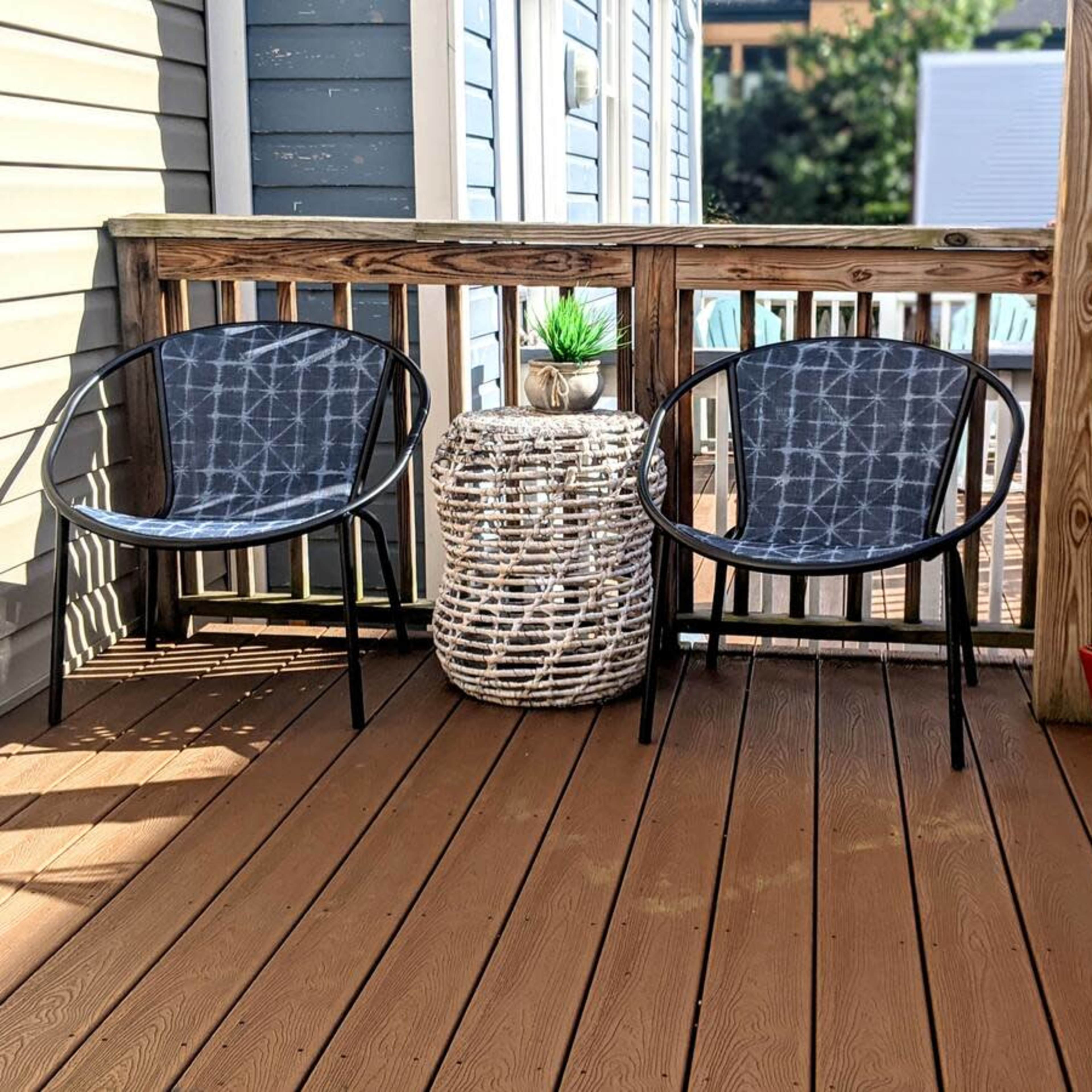 Two black chairs and a round woven table on a wooden deck, with a potted plant in the center and a hammock hanging nearby.