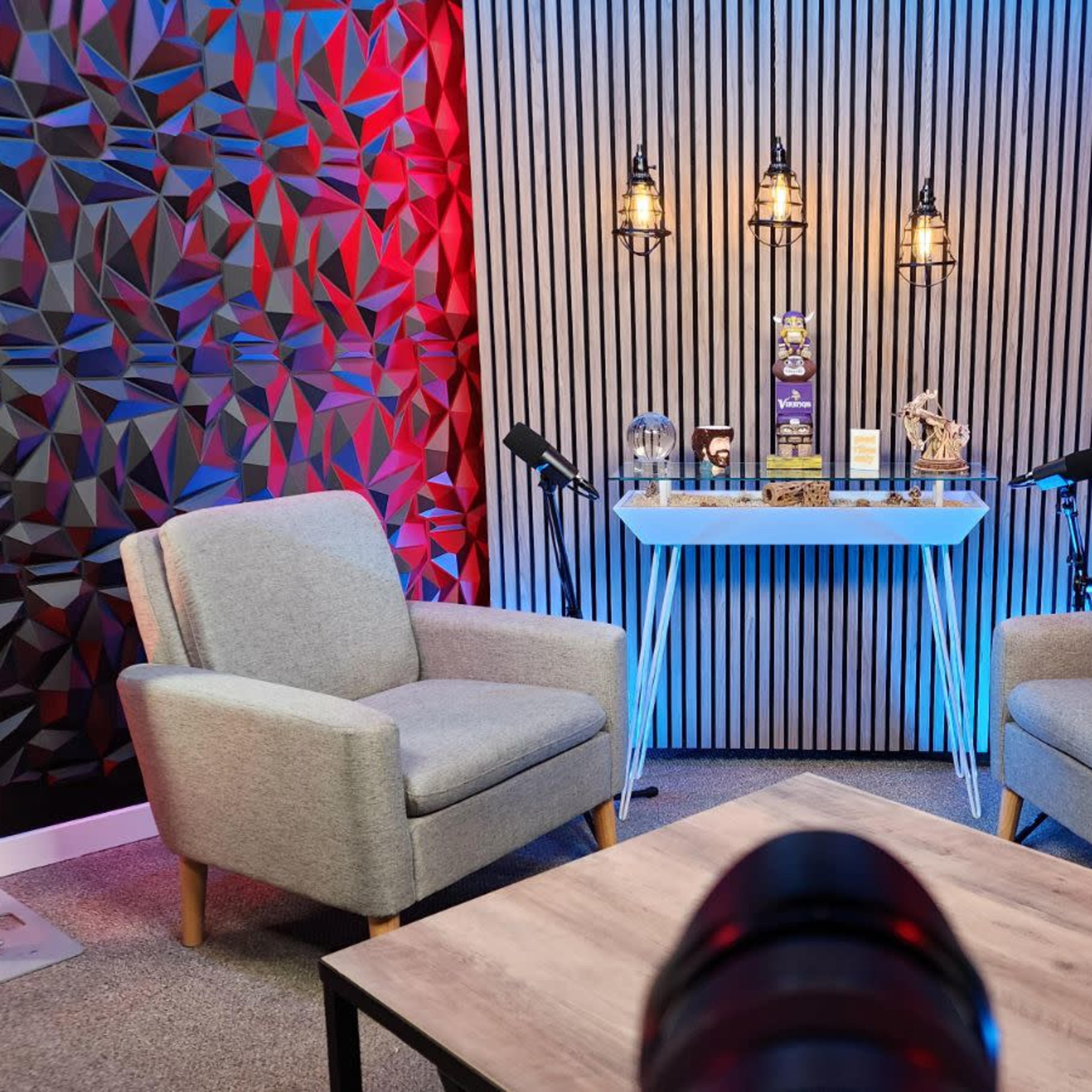A modern podcasting studio featuring two gray chairs, a wooden table, and a backdrop with patterned walls illuminated by red and blue lights.