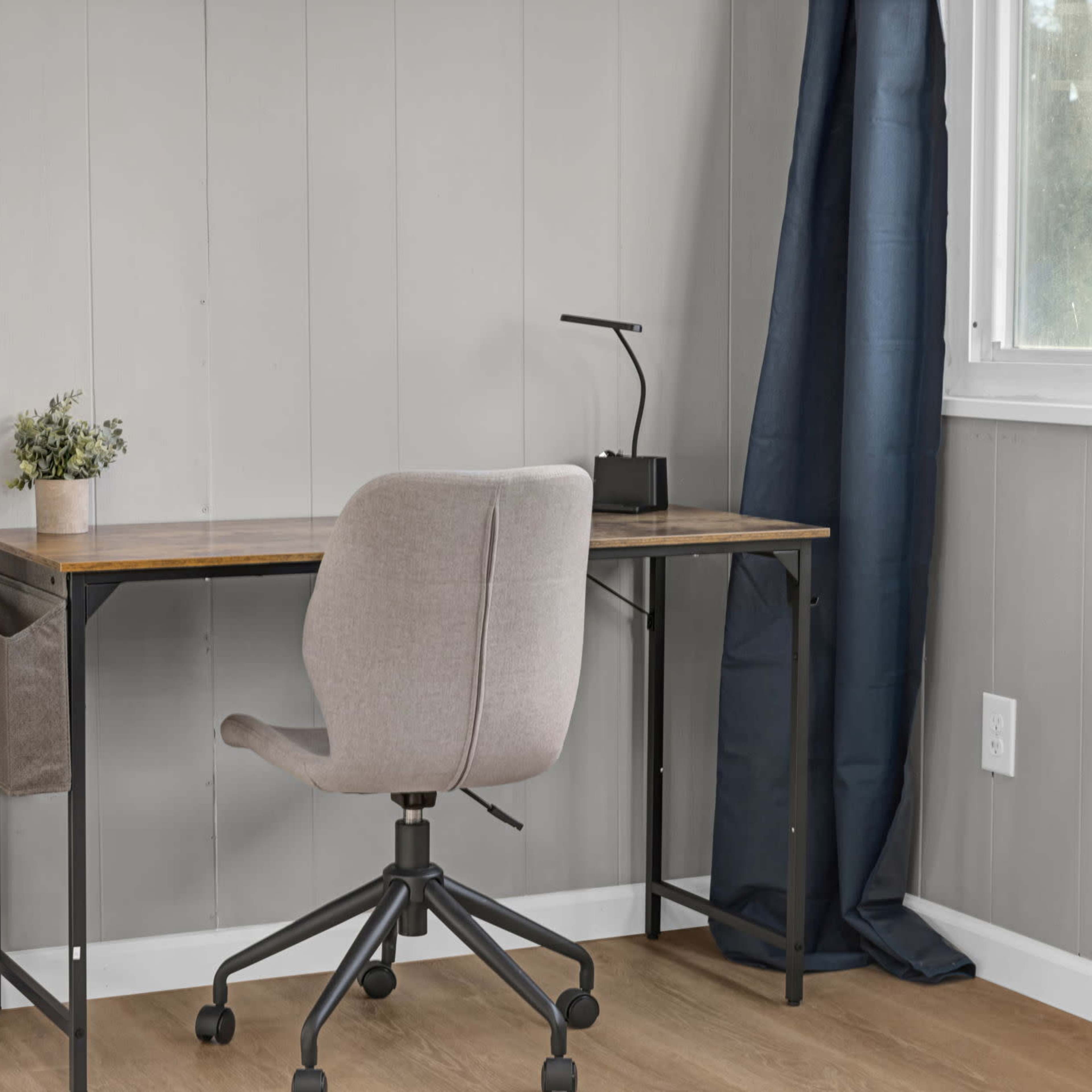 A minimalist workspace with a wooden desk, a gray chair, and a small plant on the desk, complemented by dark curtains and a lamp.
